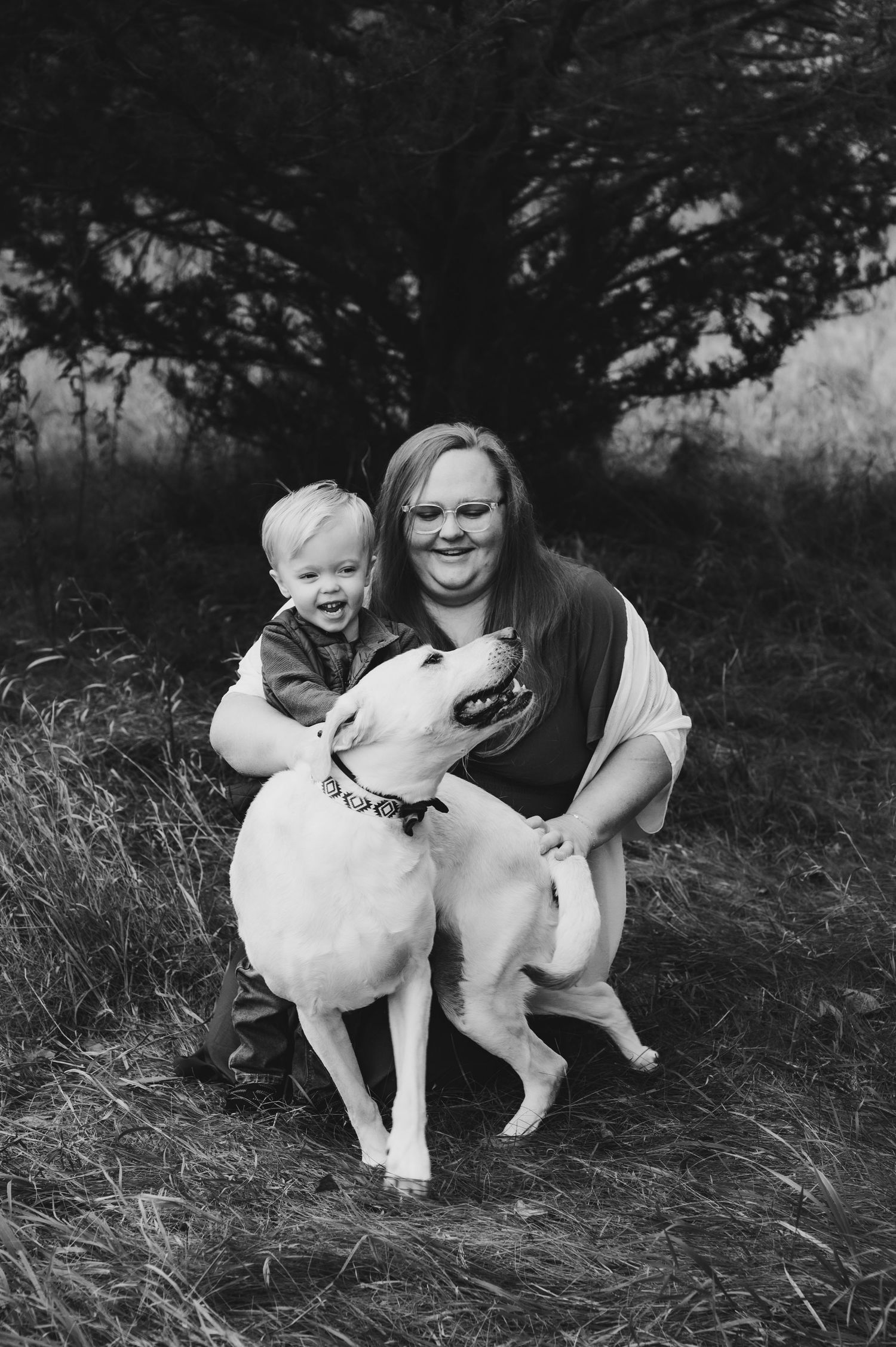 A black and white portrait captures a joyful moment outdoors with a white Labrador and family on grassy terrain.