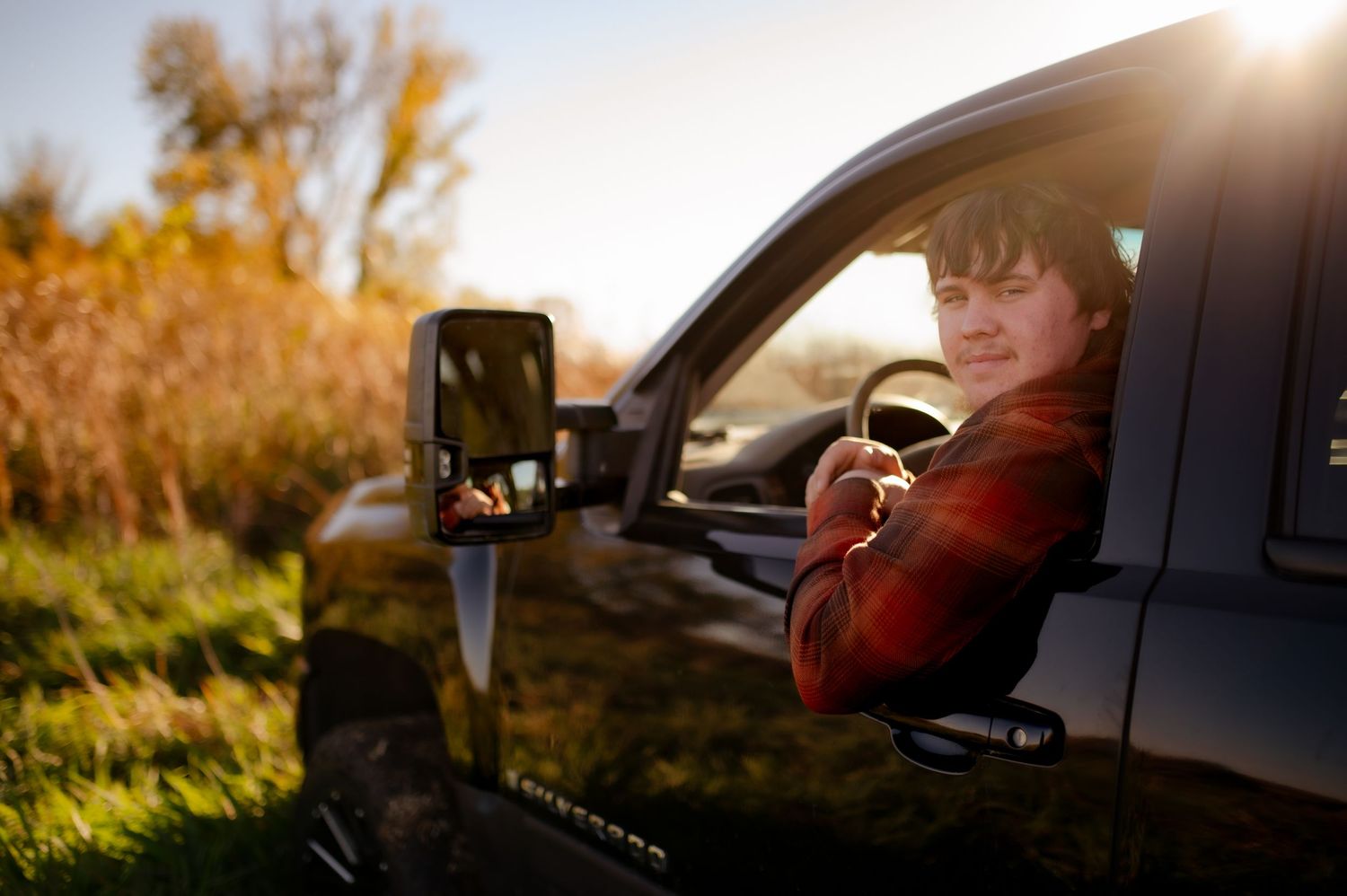 Person in red sweater sitting in driver's seat of car during golden hour with sun flare in background.