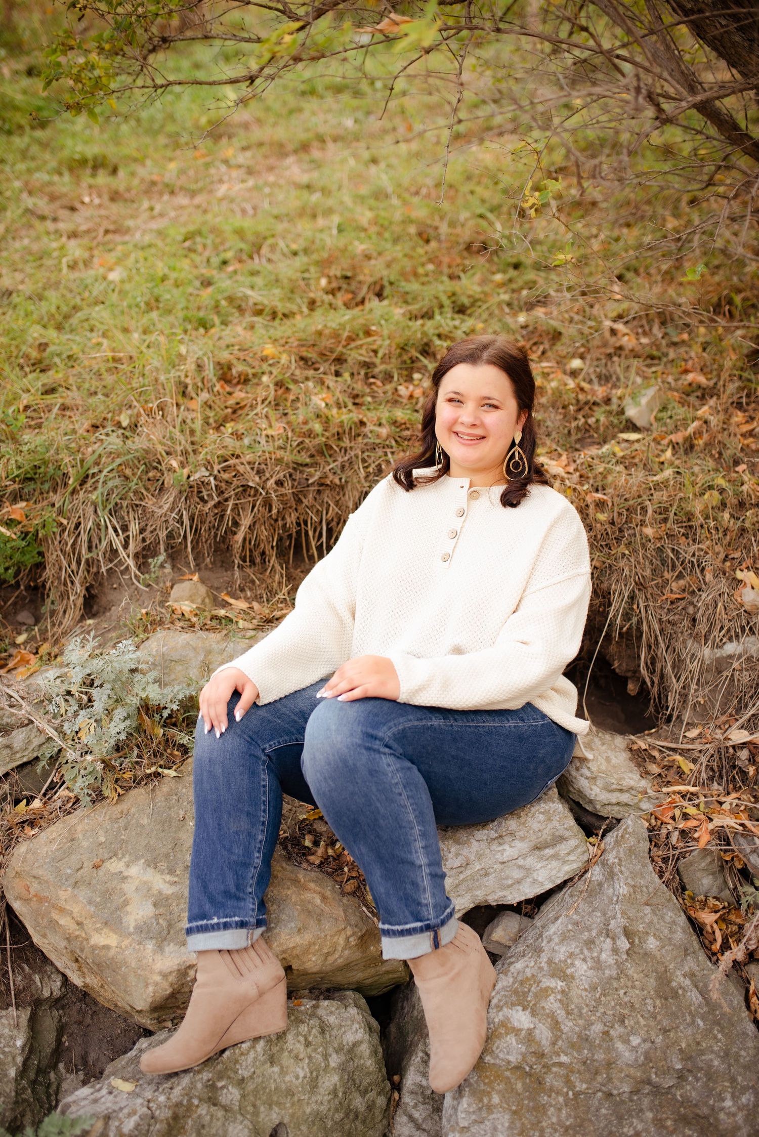 A person in a white sweater and jeans sits casually on large rocks in an outdoor autumn setting.