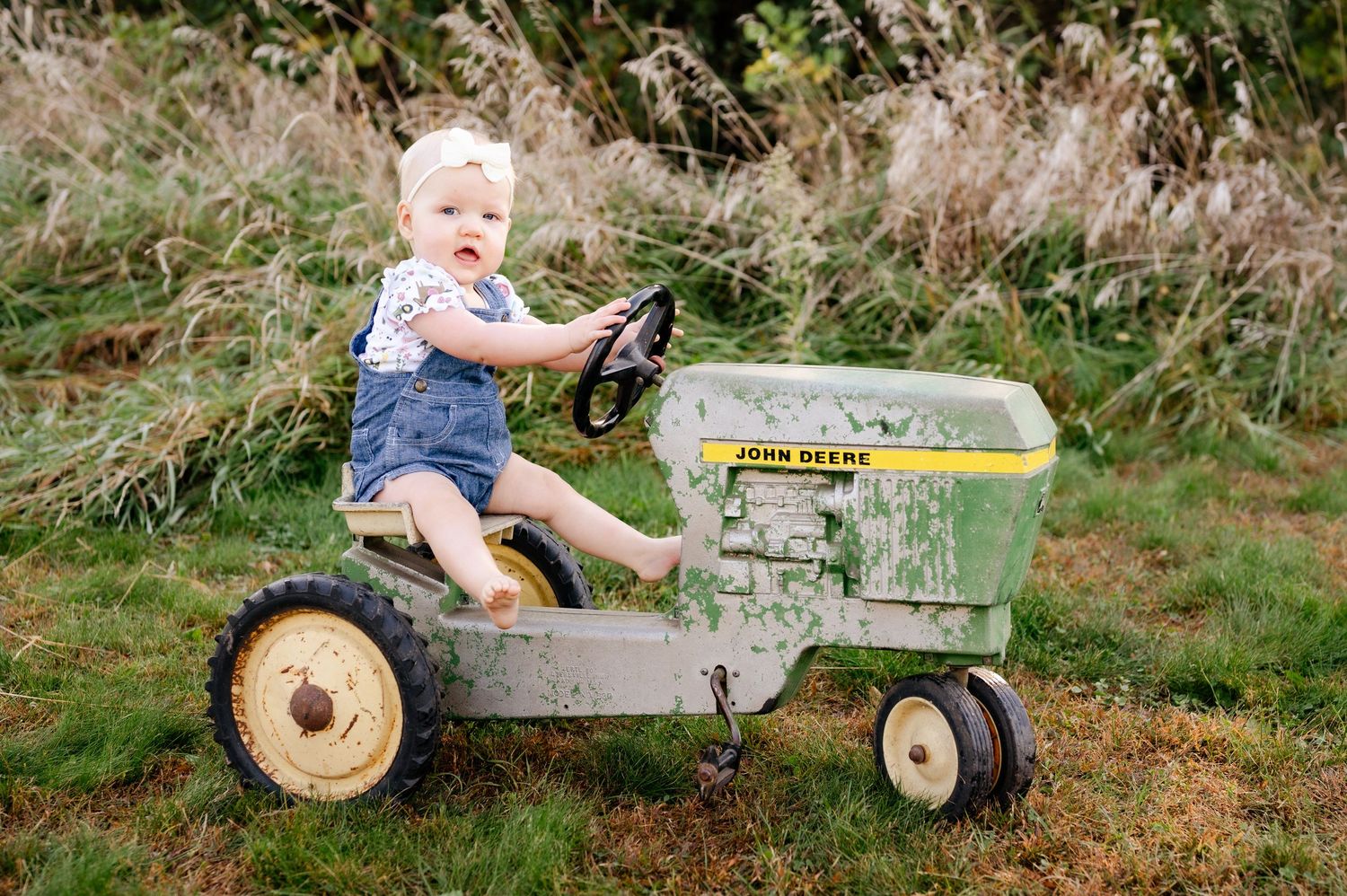 Small child rides vintage green John Deere pedal tractor in grassy outdoor setting.