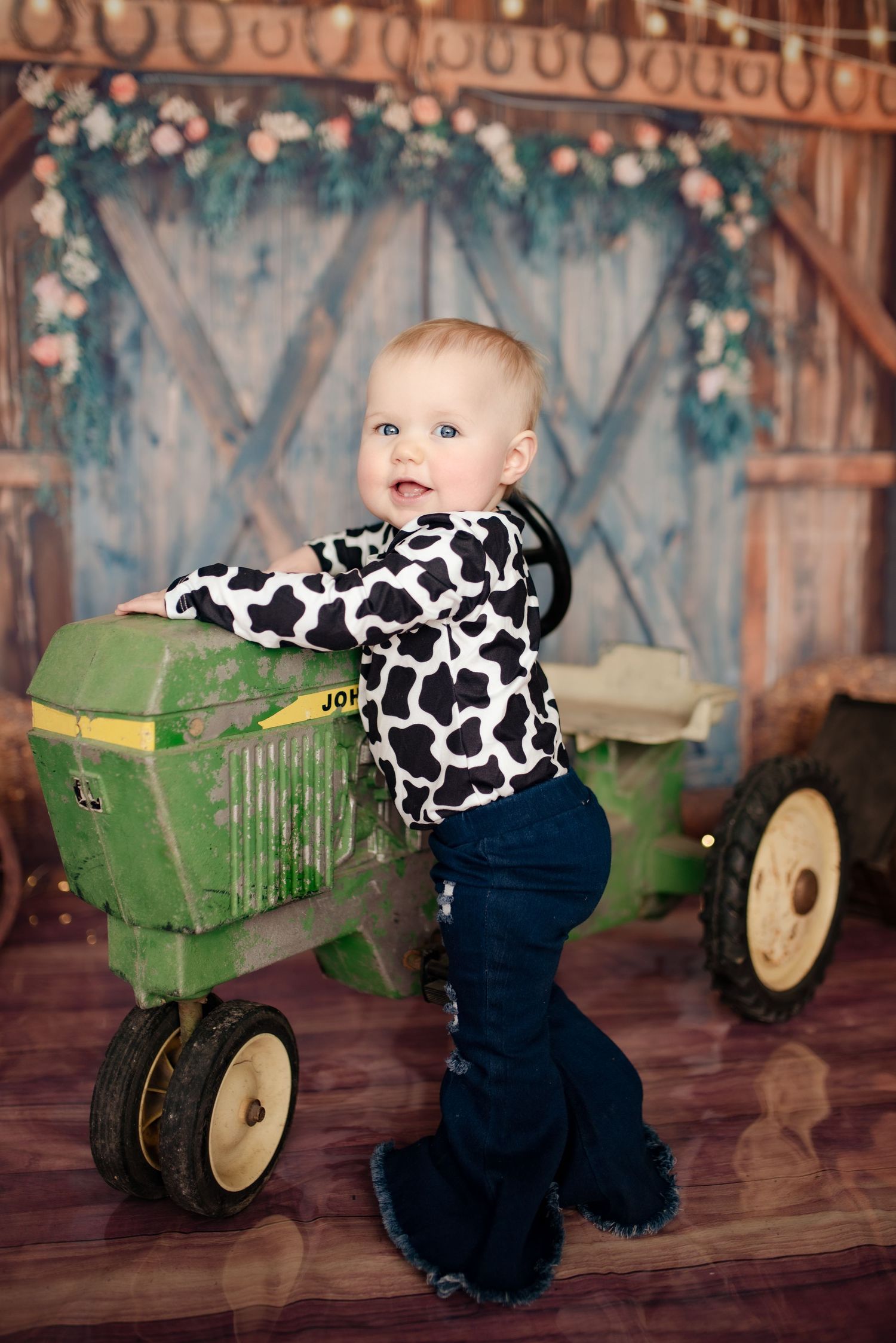 Small child in cow print top plays with vintage green toy tractor against rustic barn backdrop with floral garland.