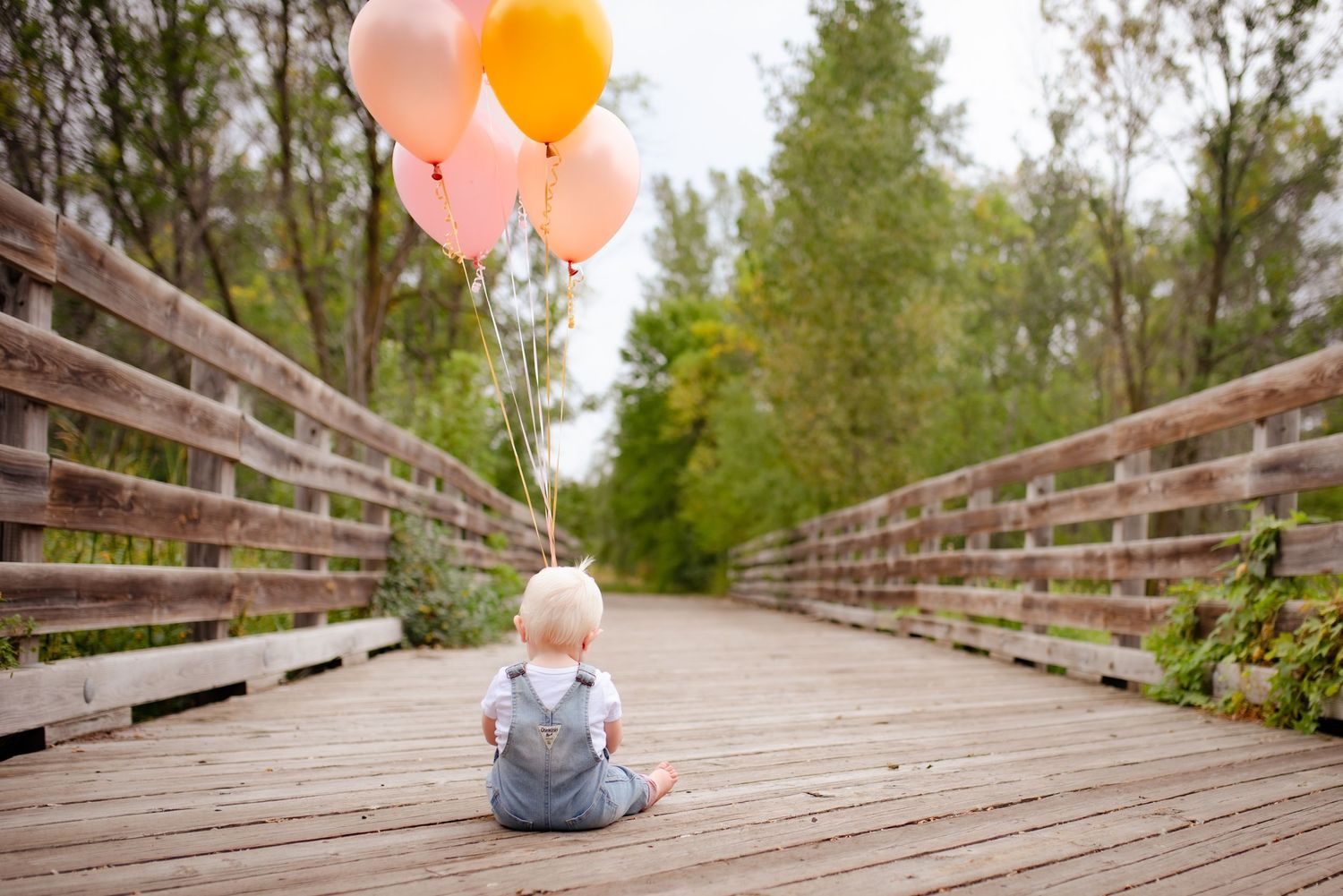Small child sits on wooden boardwalk holding pastel balloons with trees in background.