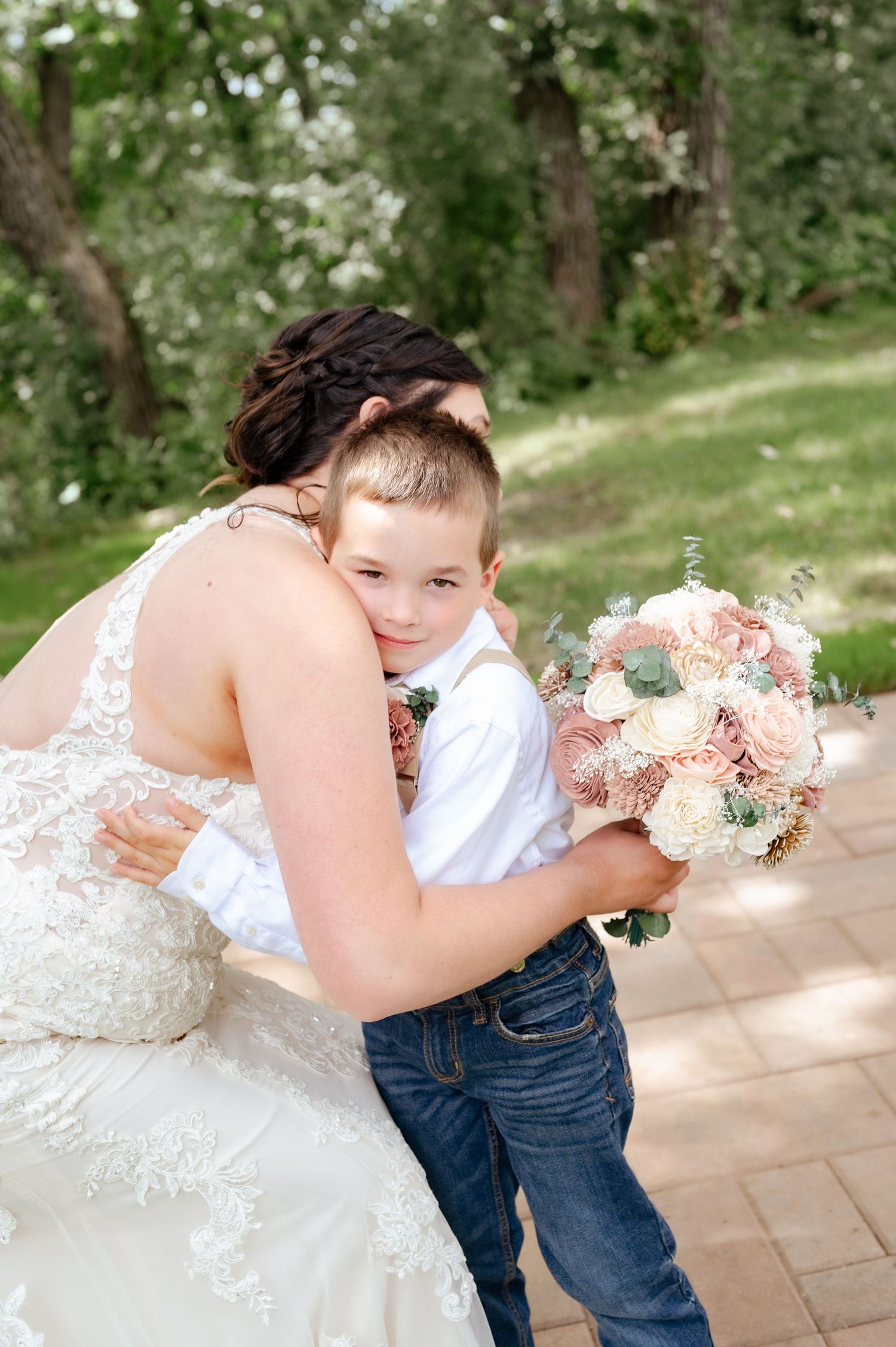 Tender moment between bride in lace dress holding bouquet and young boy in formal attire outdoors.