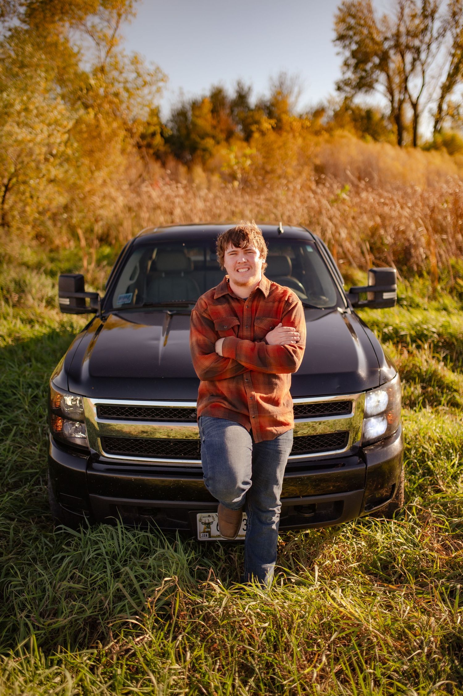 Black Chevrolet car parked in tall grass during golden hour with autumn foliage in background.