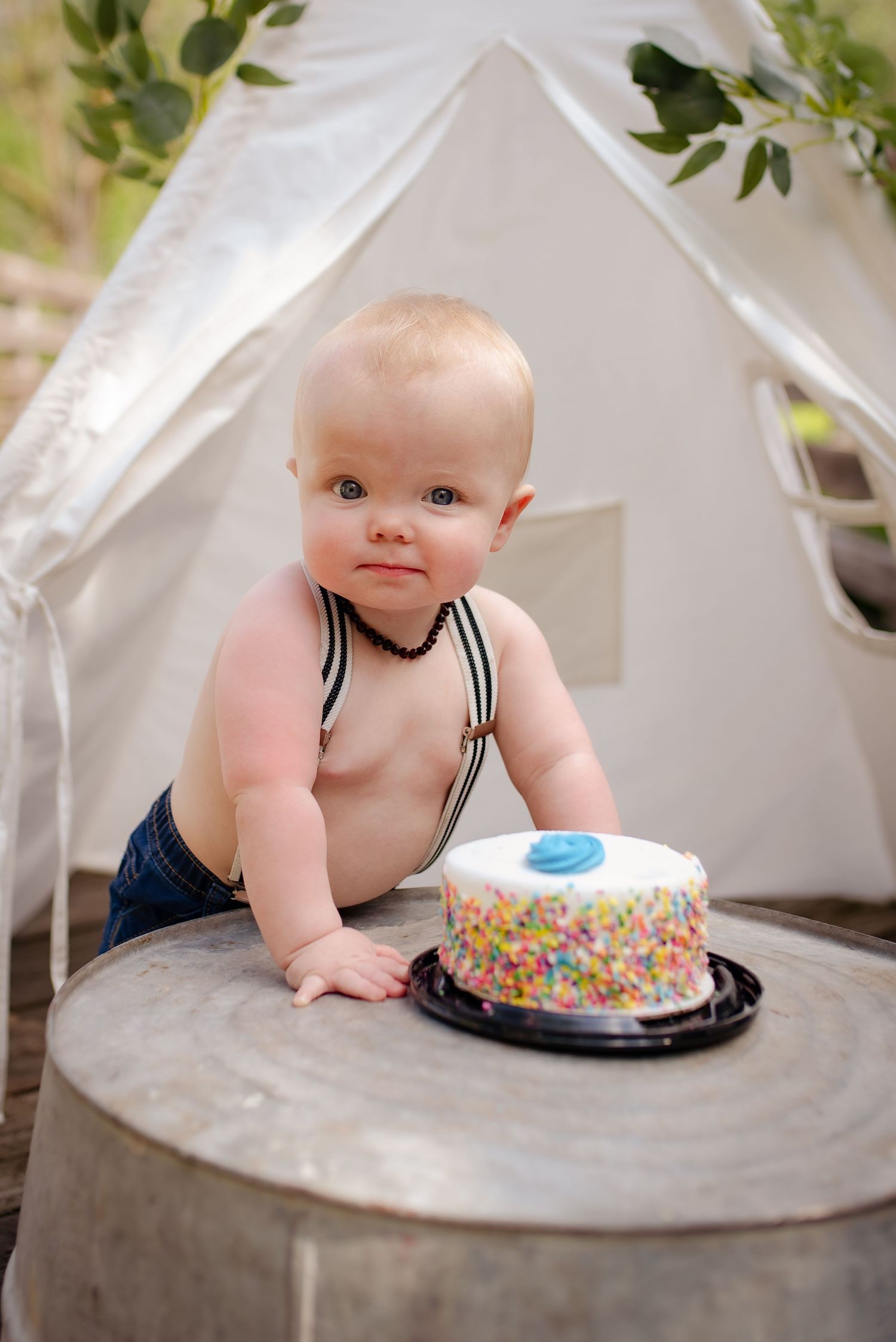 A baby in navy suspenders sits with a sprinkle-covered birthday cake inside a white tent.