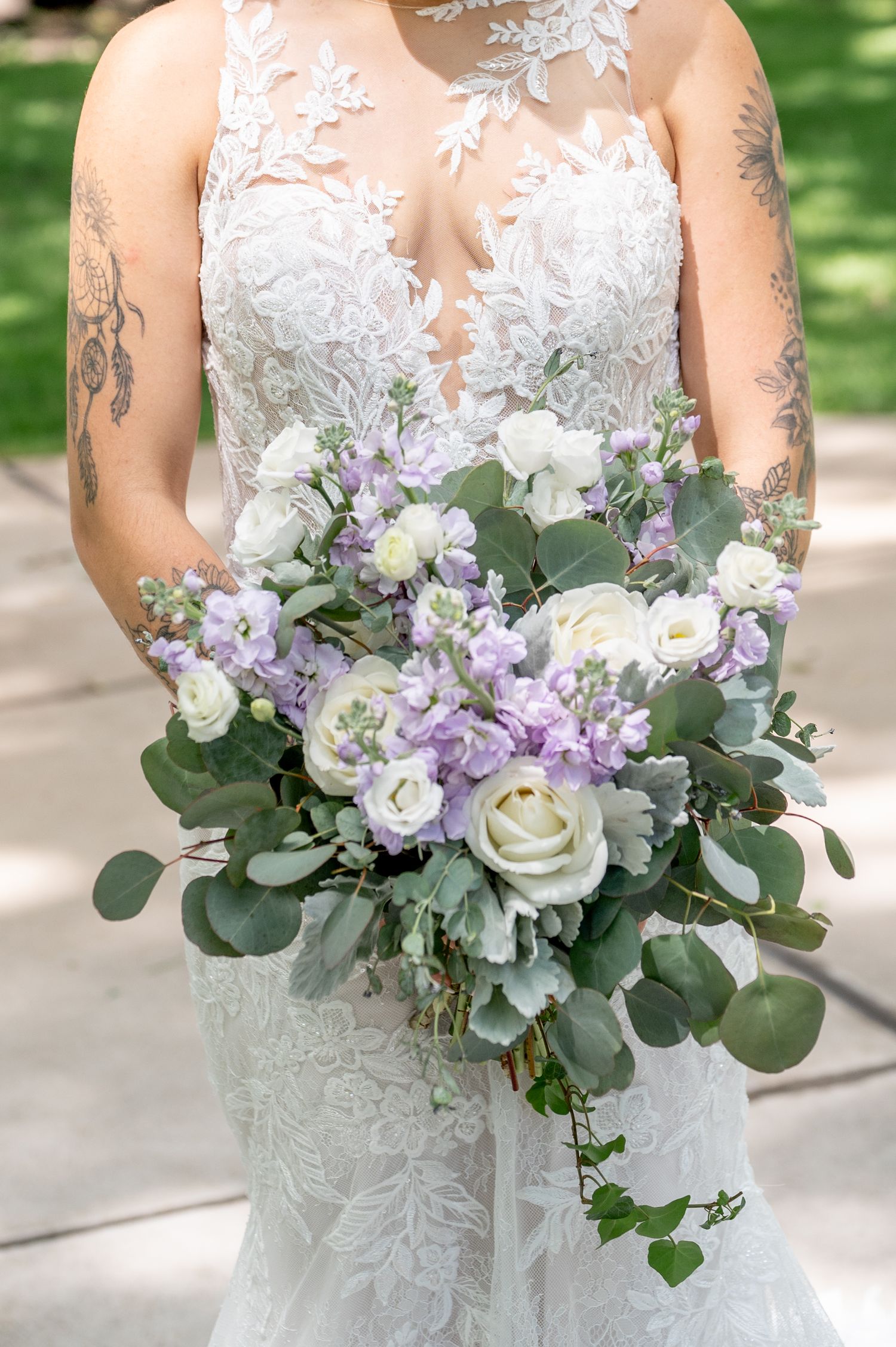 Close-up of a stunning bridal bouquet with white roses, purple flowers, and eucalyptus against a lace wedding dress.