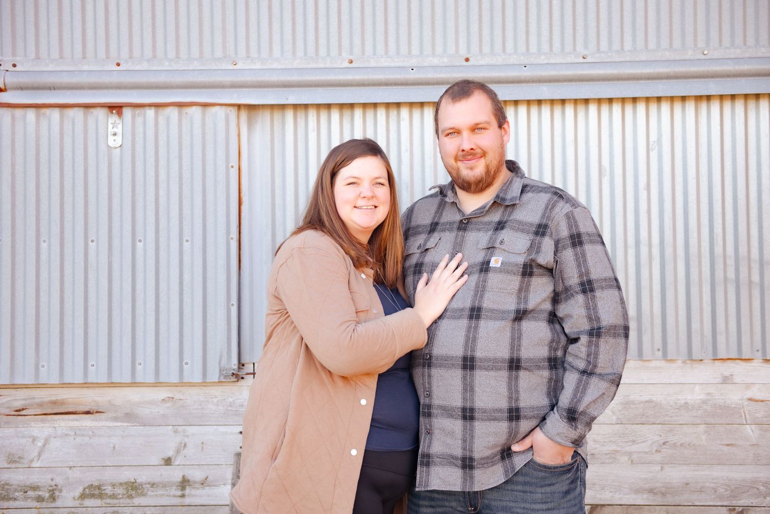 Couple poses together against industrial metal wall, wearing casual winter attire.