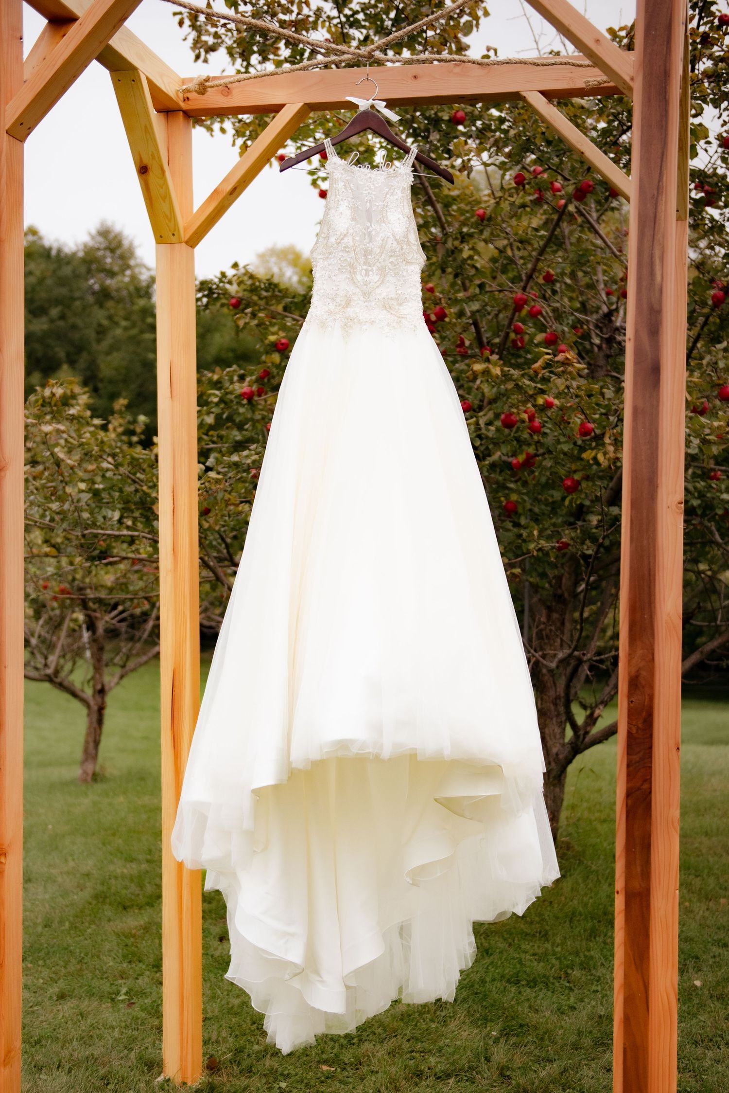 Elegant white wedding dress hanging from wooden pergola in garden setting with sunlit trees in background.