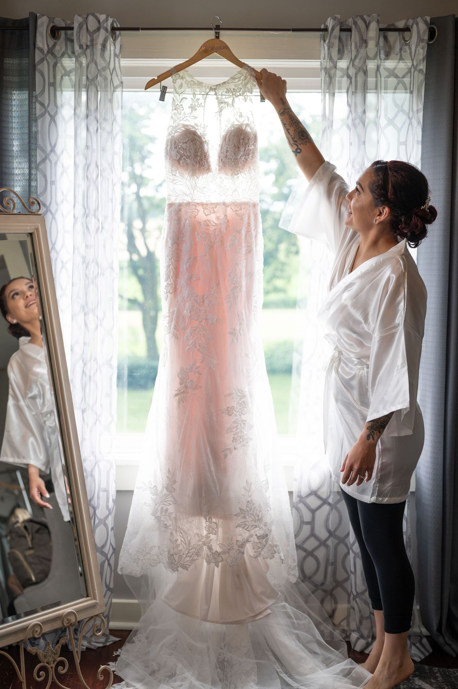 A beautiful blush wedding dress with lace details hangs in front of a sunlit window with sheer curtains.