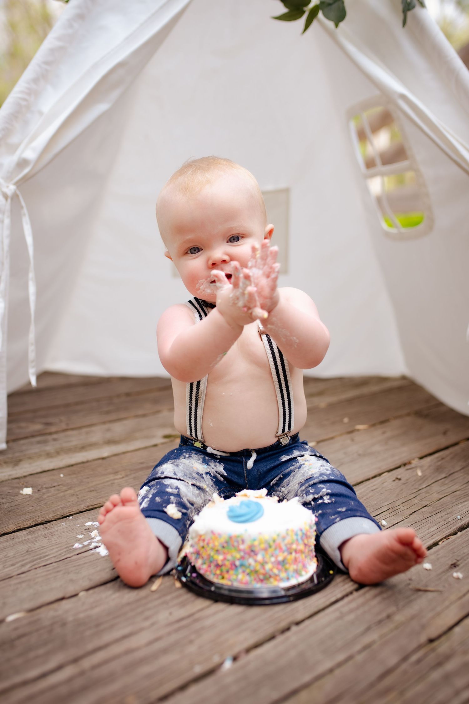 A joyful baby smashes into a white and blue birthday cake while sitting under a white tent.