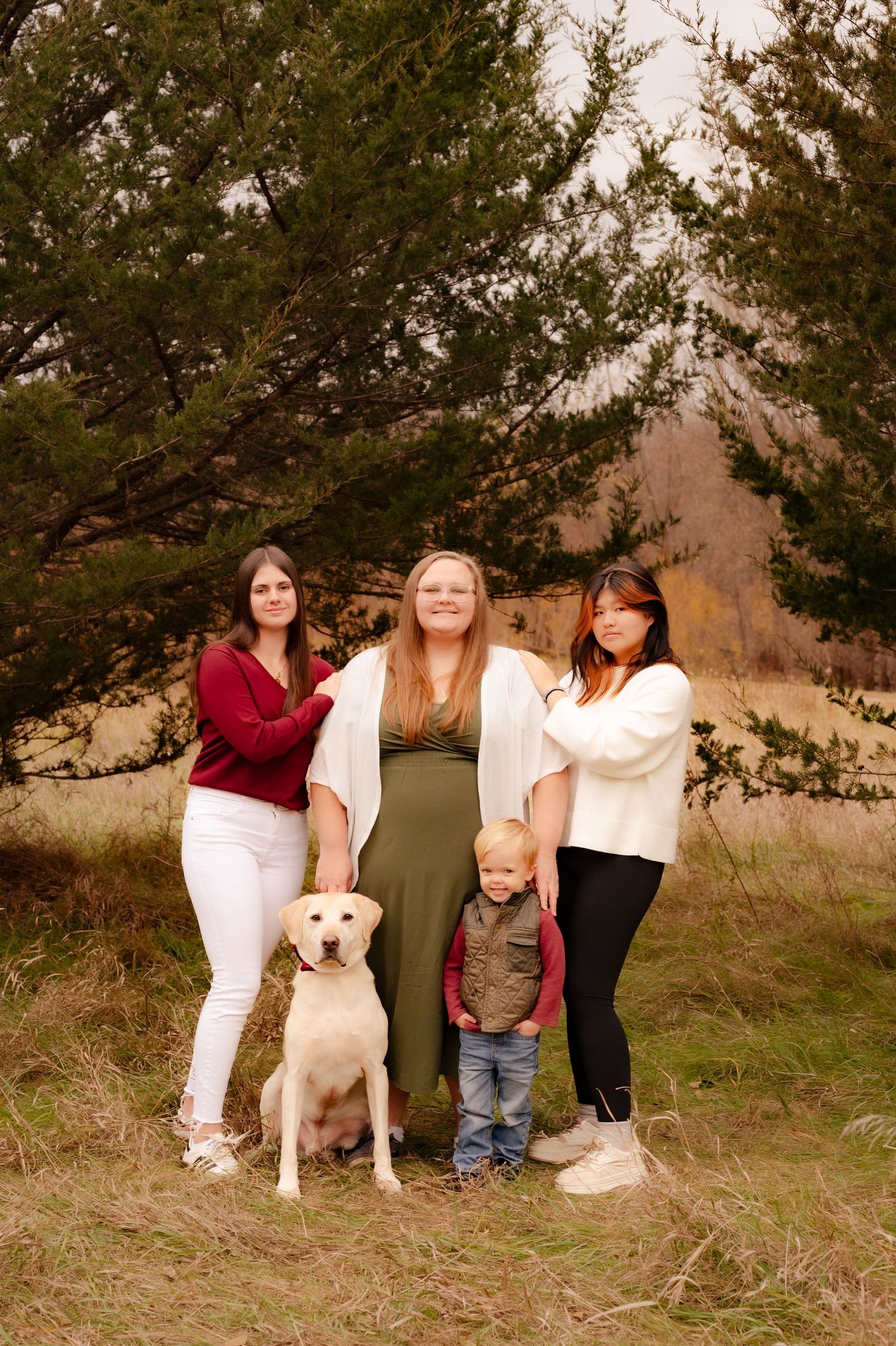 A group poses together outdoors near evergreen trees with a white dog and small child on a fall day.
