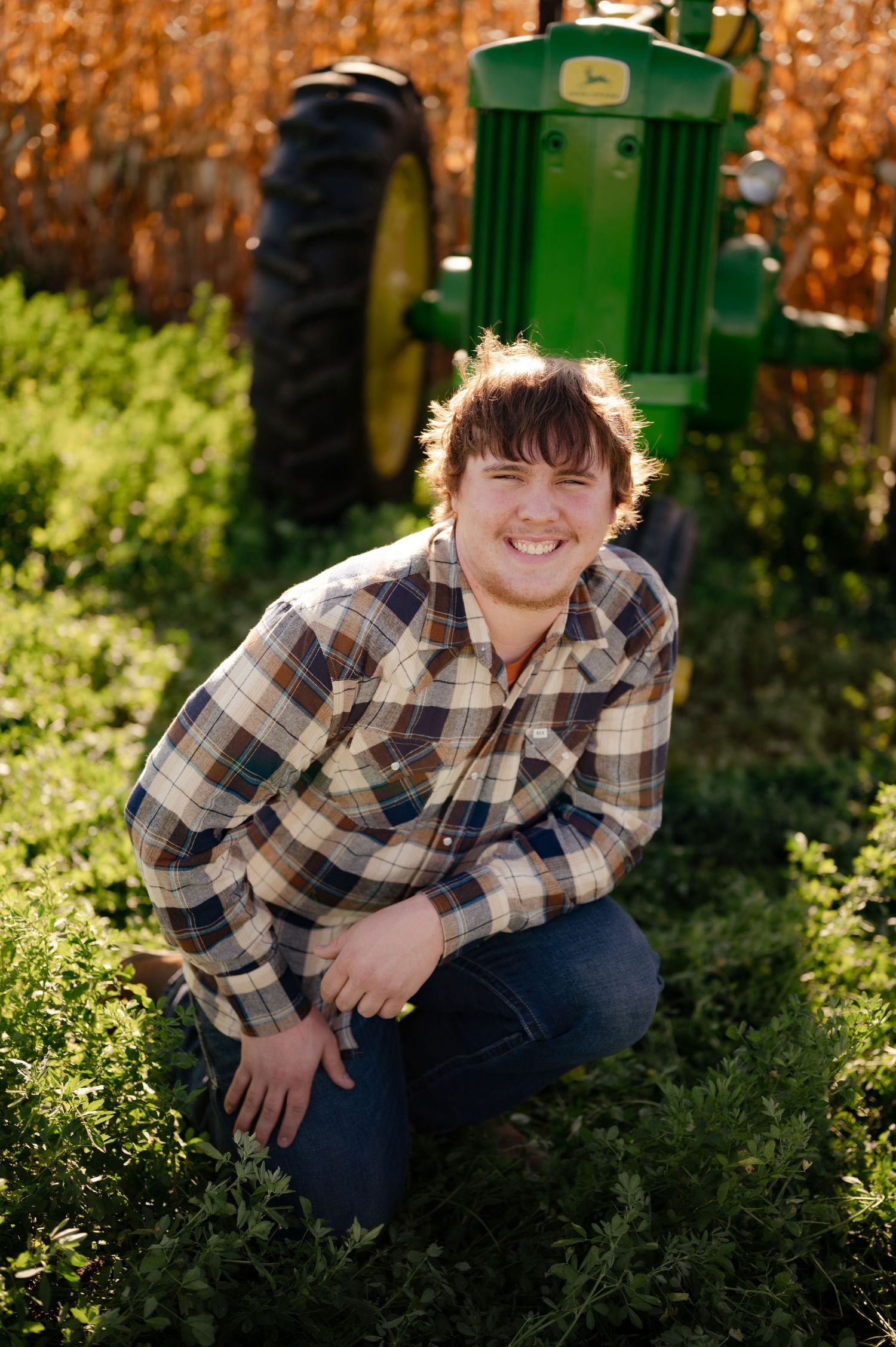 Person in plaid shirt poses casually in front of green tractor with golden cornfield in background.