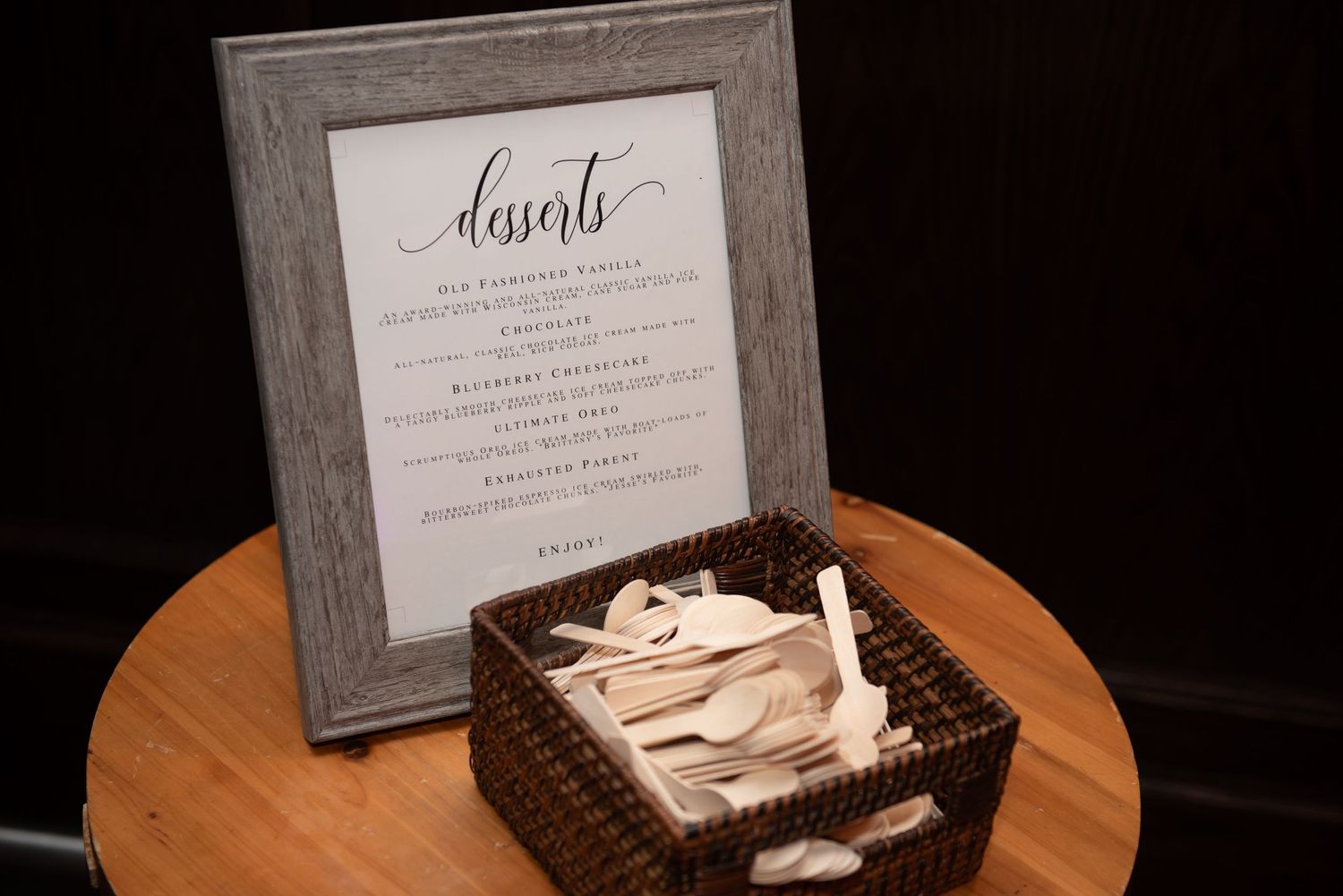 A framed wedding dessert menu sits beside a woven basket on a wooden table.