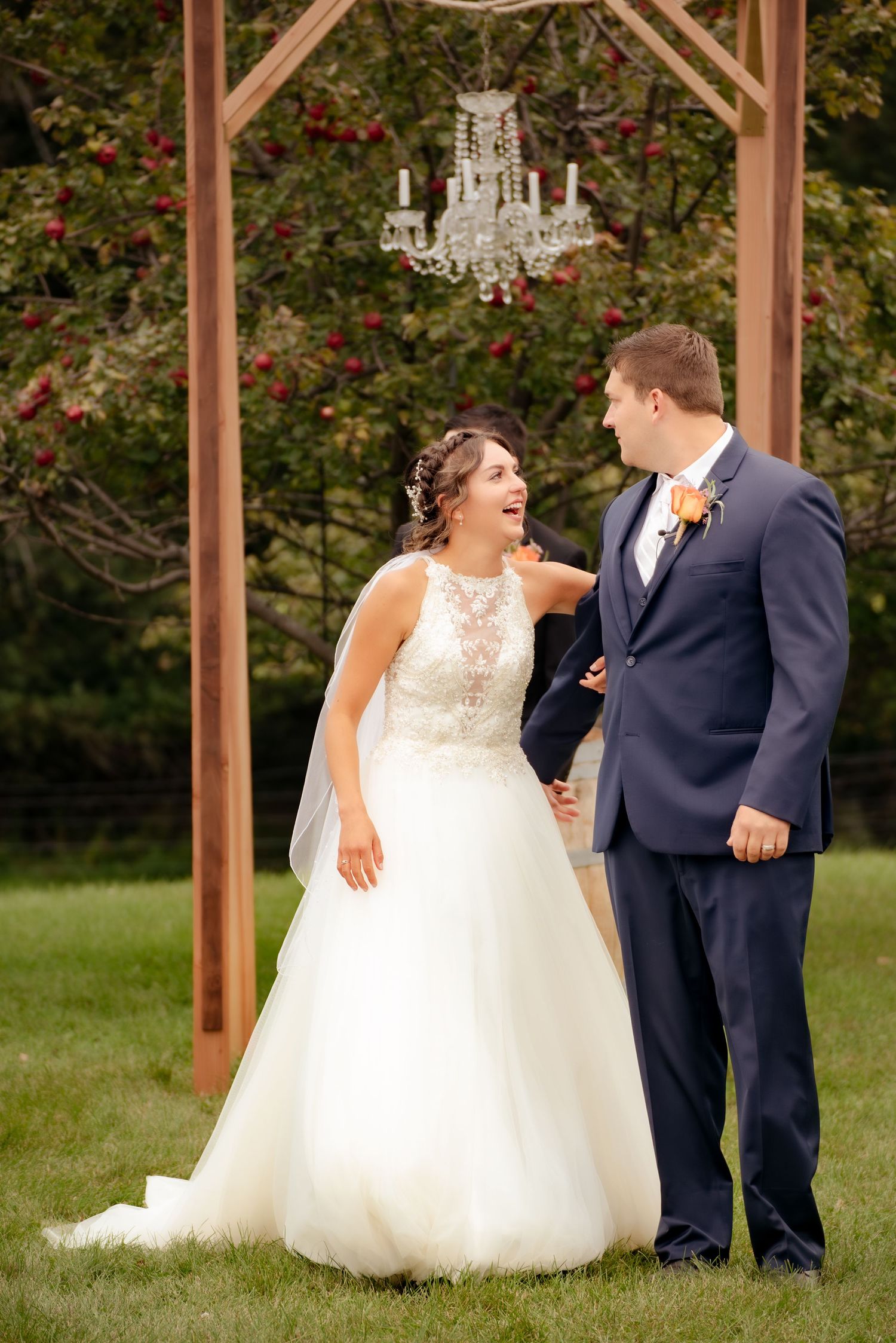 Couple shares a joyful moment under a crystal chandelier and wooden arch during their outdoor wedding.