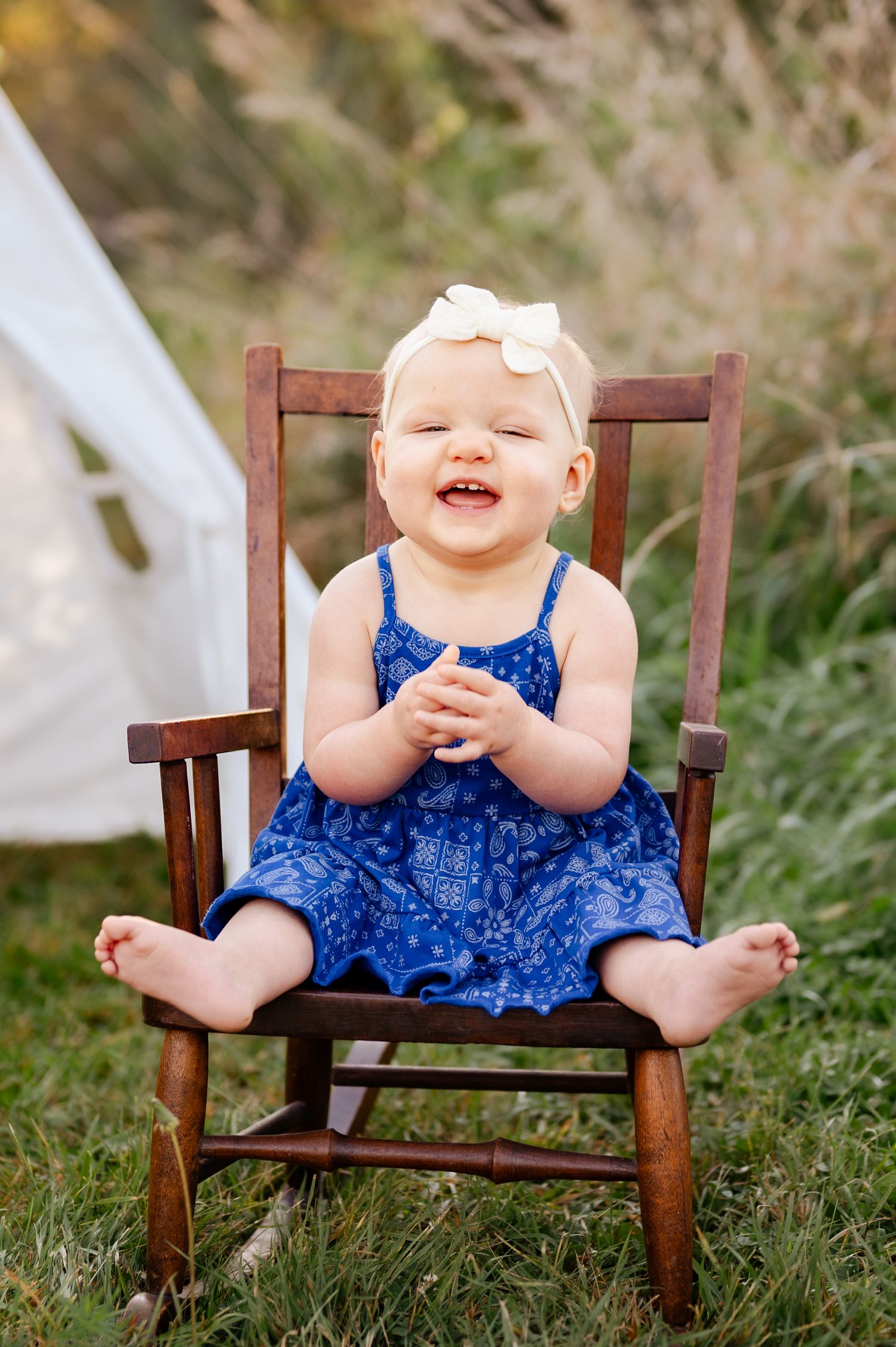 A joyful toddler wearing a blue dress and white bow sits in a wooden rocking chair outdoors.