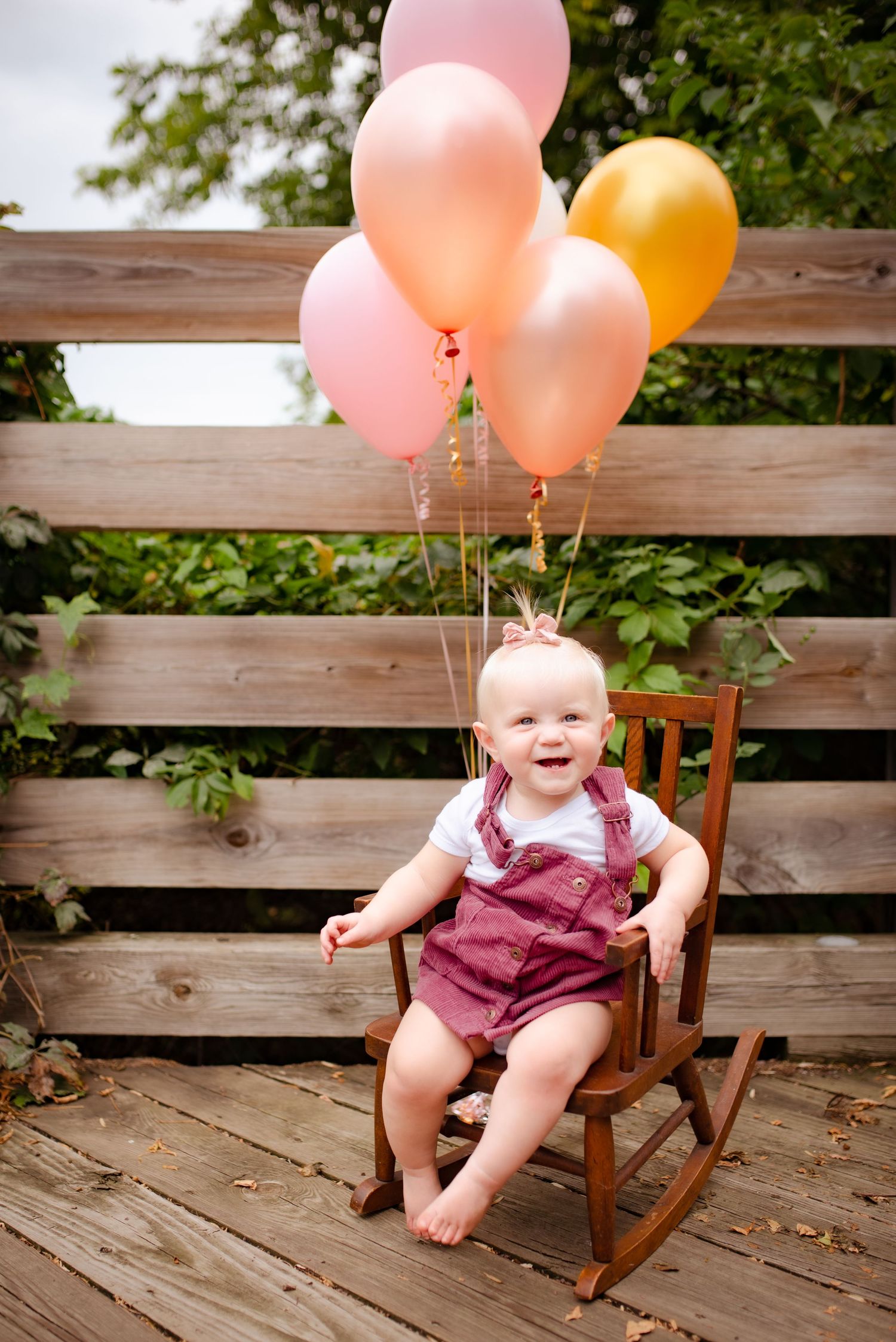 Happy baby in red overalls sits in rocking chair with pink and orange balloons.