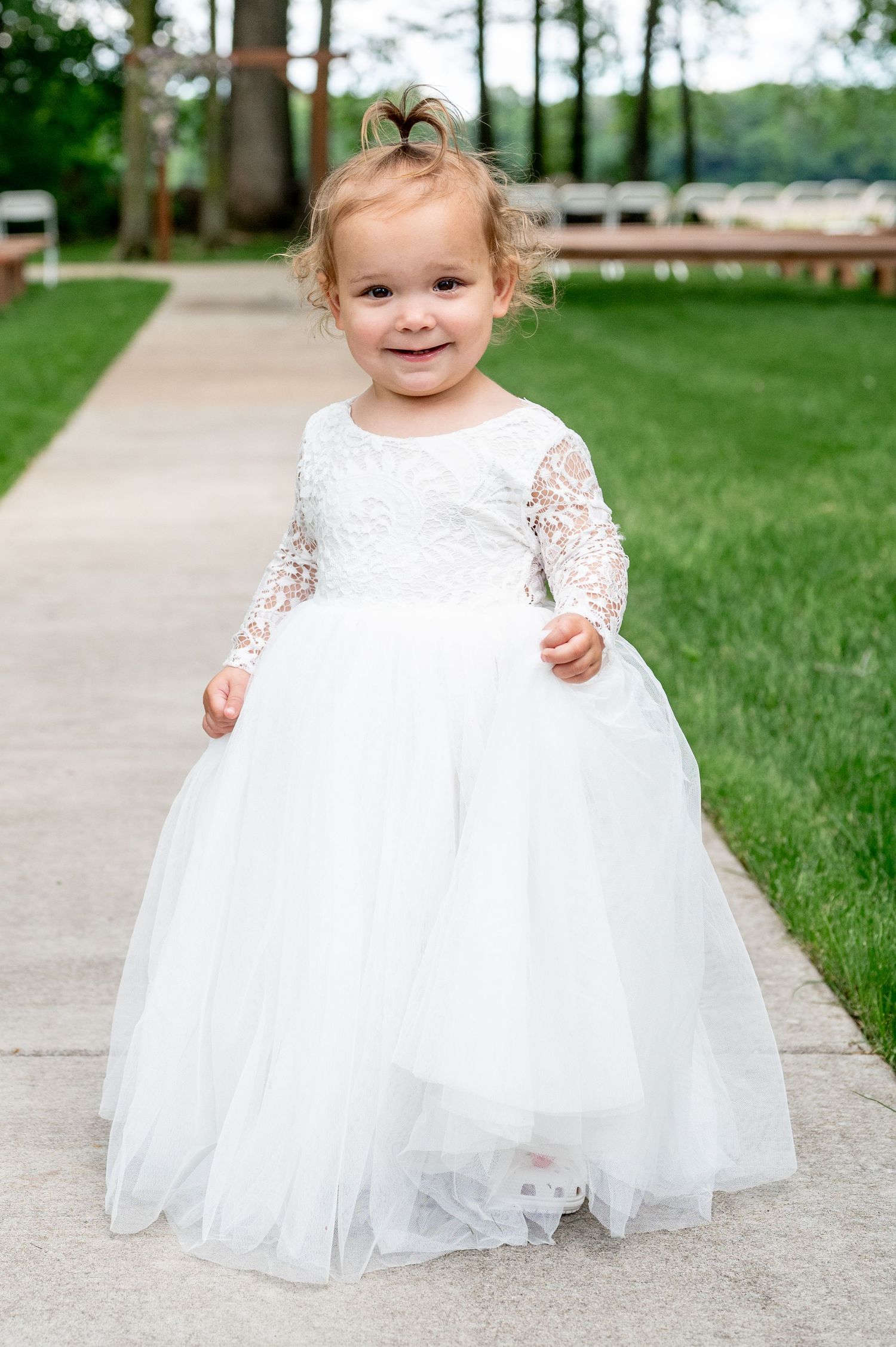 Smiling flower girl wearing white lace and tulle dress poses on outdoor pathway.