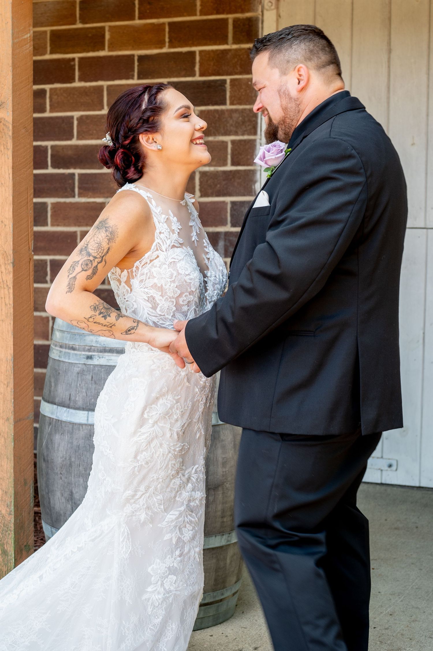 Newlyweds share a joyful moment together by a rustic brick wall and wooden barrel.