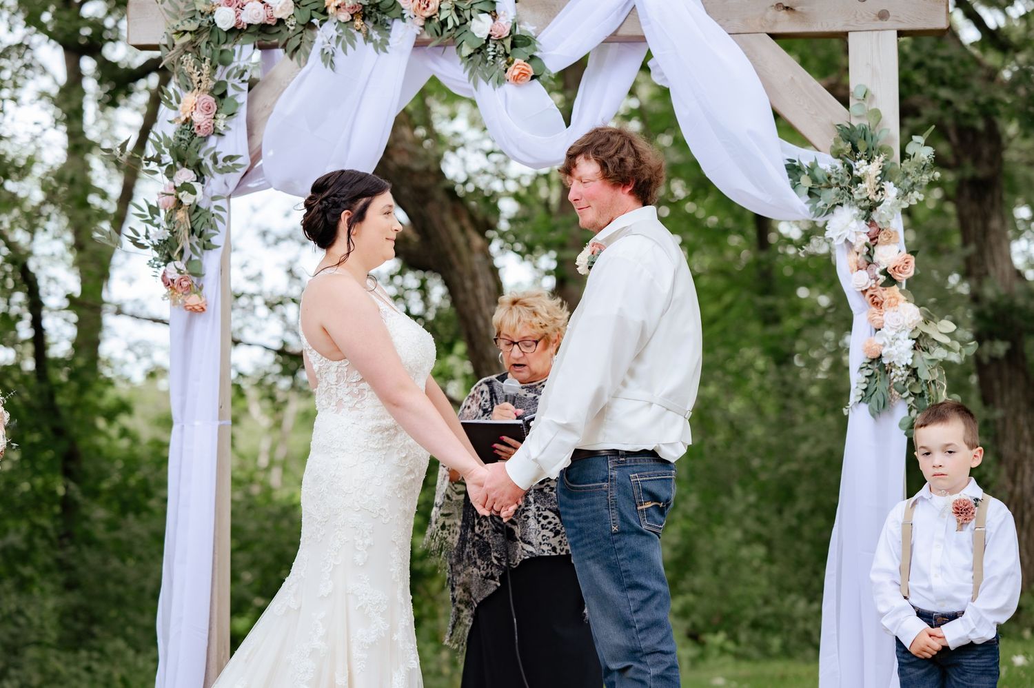 Wedding ceremony under a white arch decorated with pink flowers and delicate fabric draping.