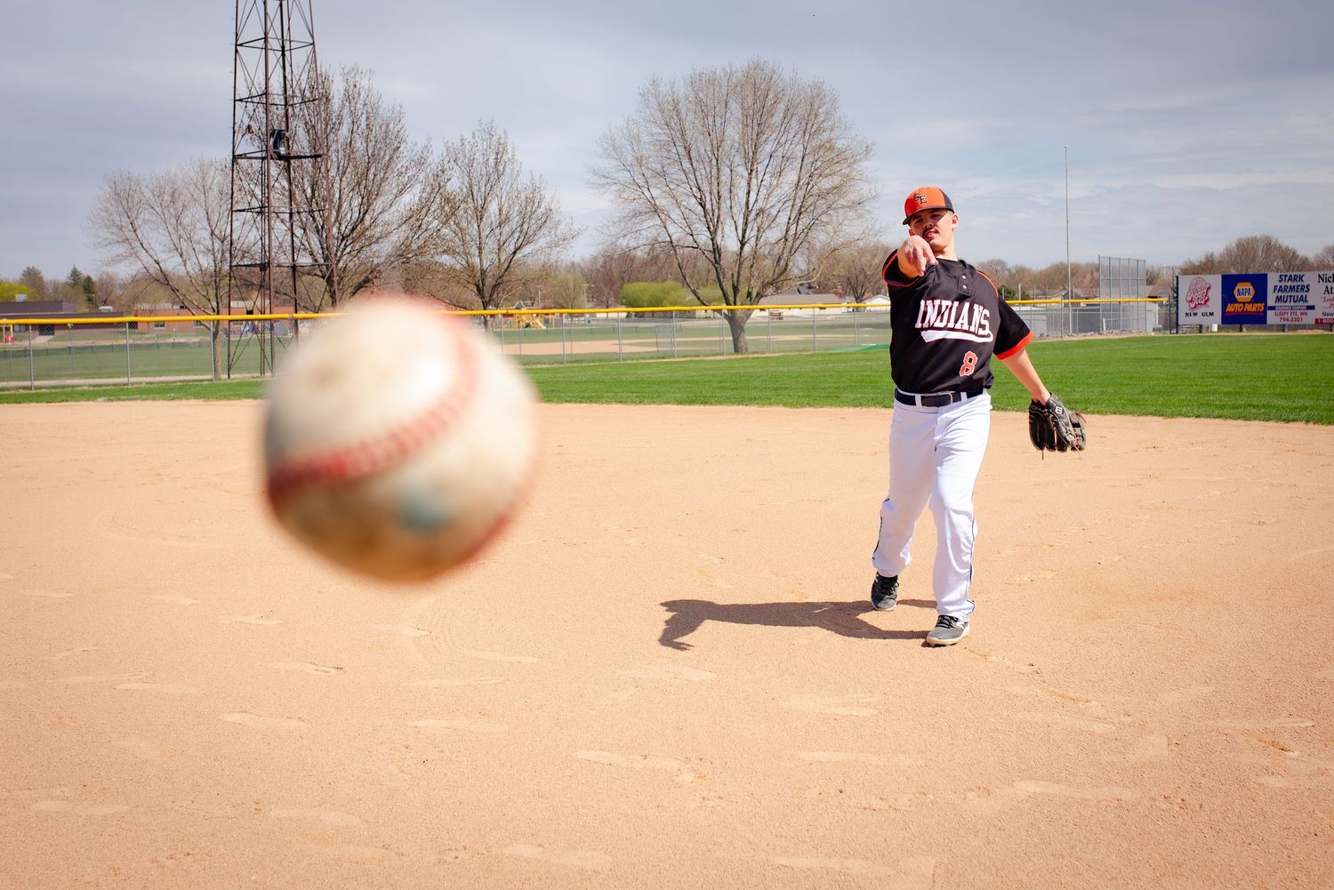 Player throws baseball on dirt field with ball in motion and bleachers in background.