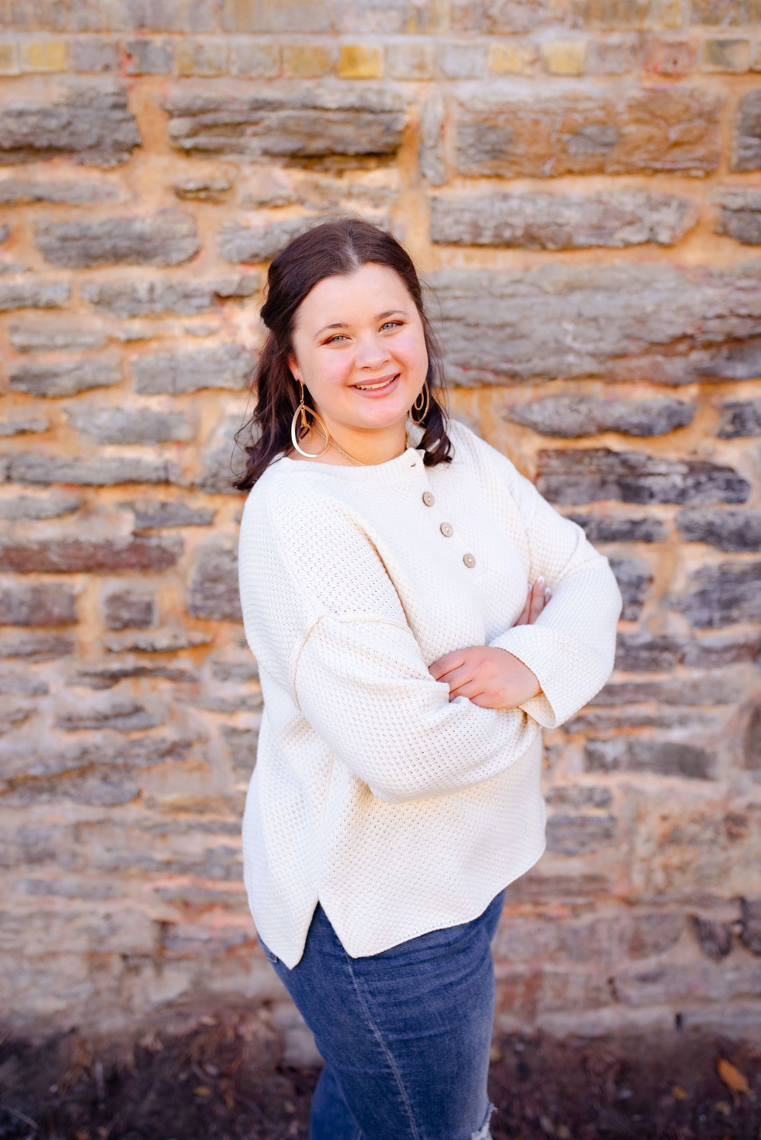 Someone smiles while posing in a white button sweater against rustic stone wall background.