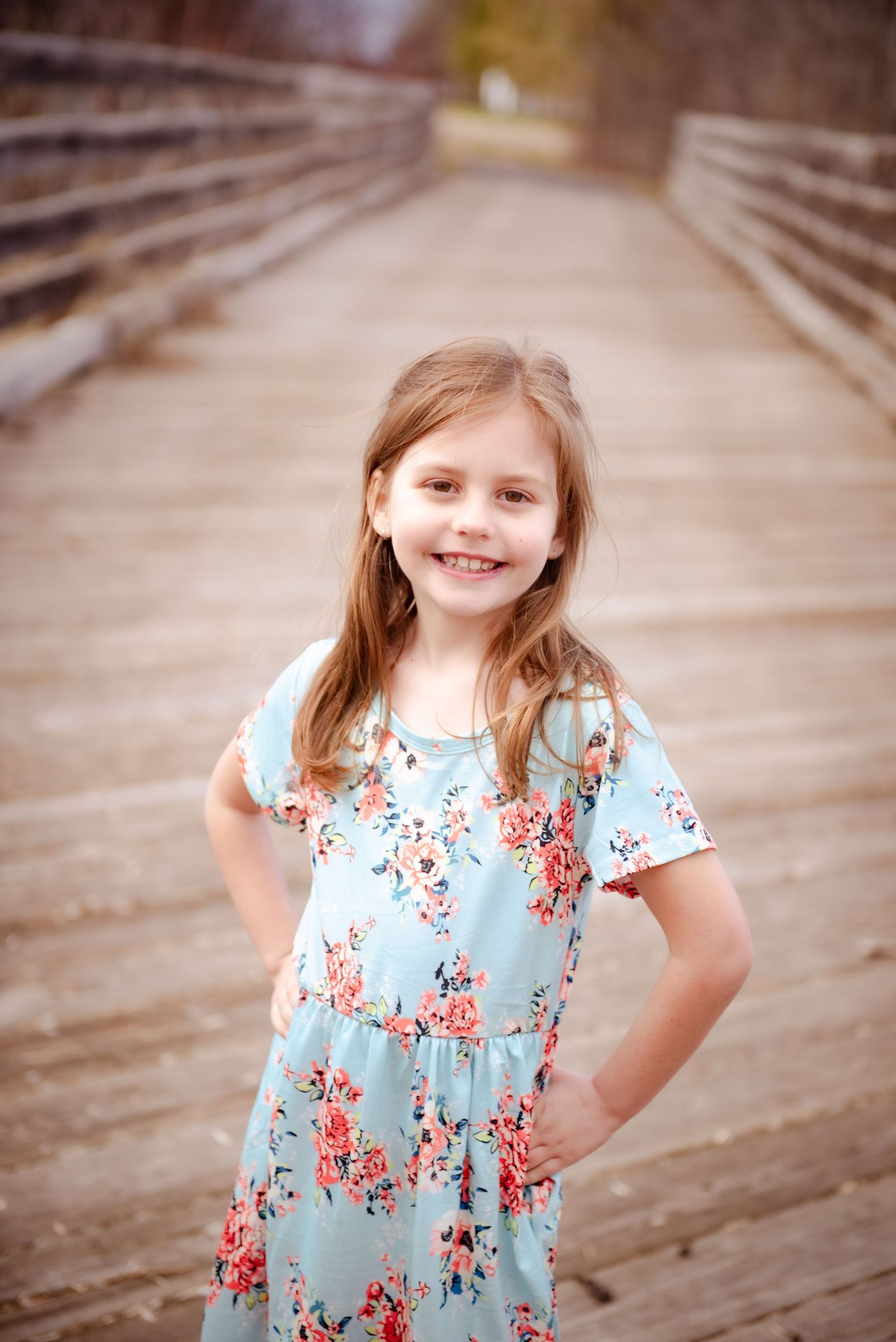 Child in light blue floral dress posing on wooden bridge.