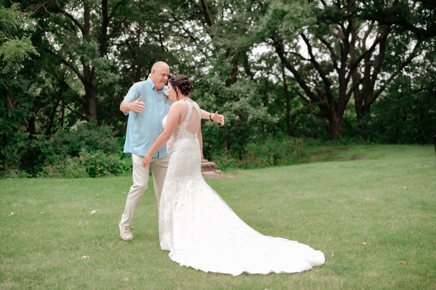 Wedding couple dancing together on green lawn surrounded by trees.