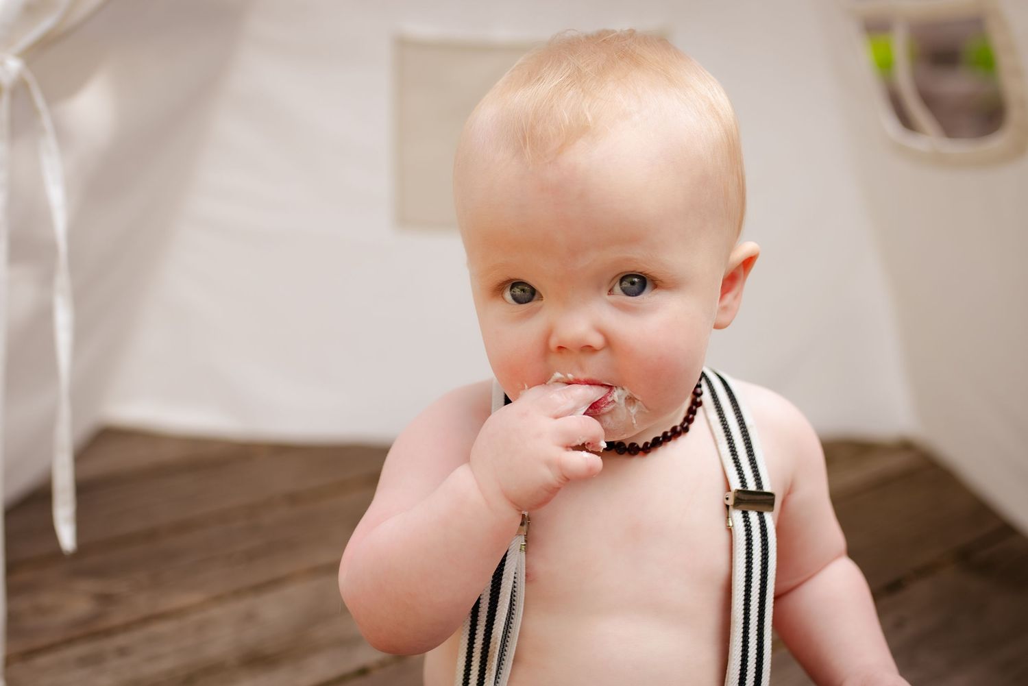 A baby in striped suspenders tastes frosting inside a white teepee tent.