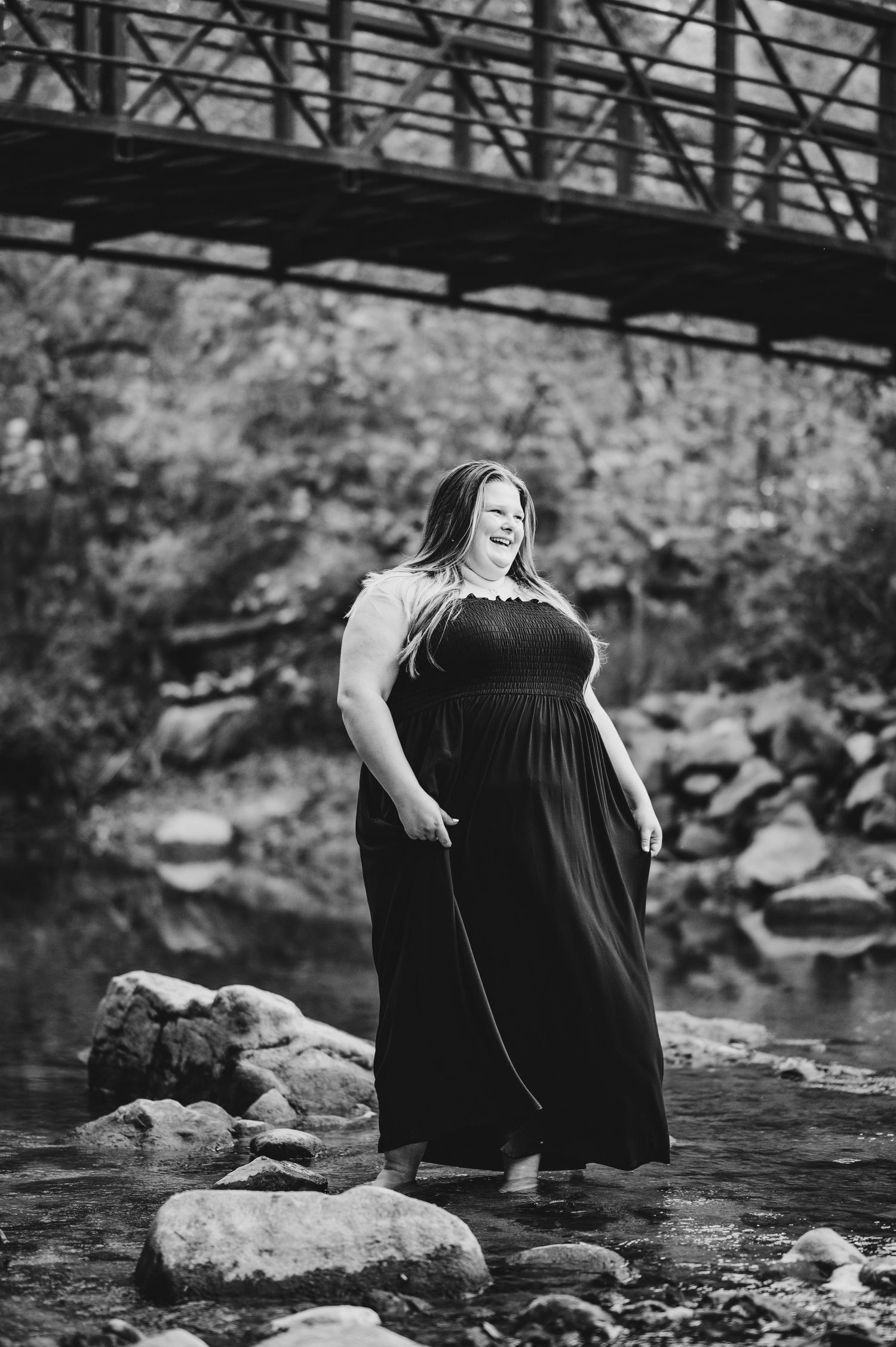 A black and white photo of a person in a long dress standing on rocks by a river under a bridge.