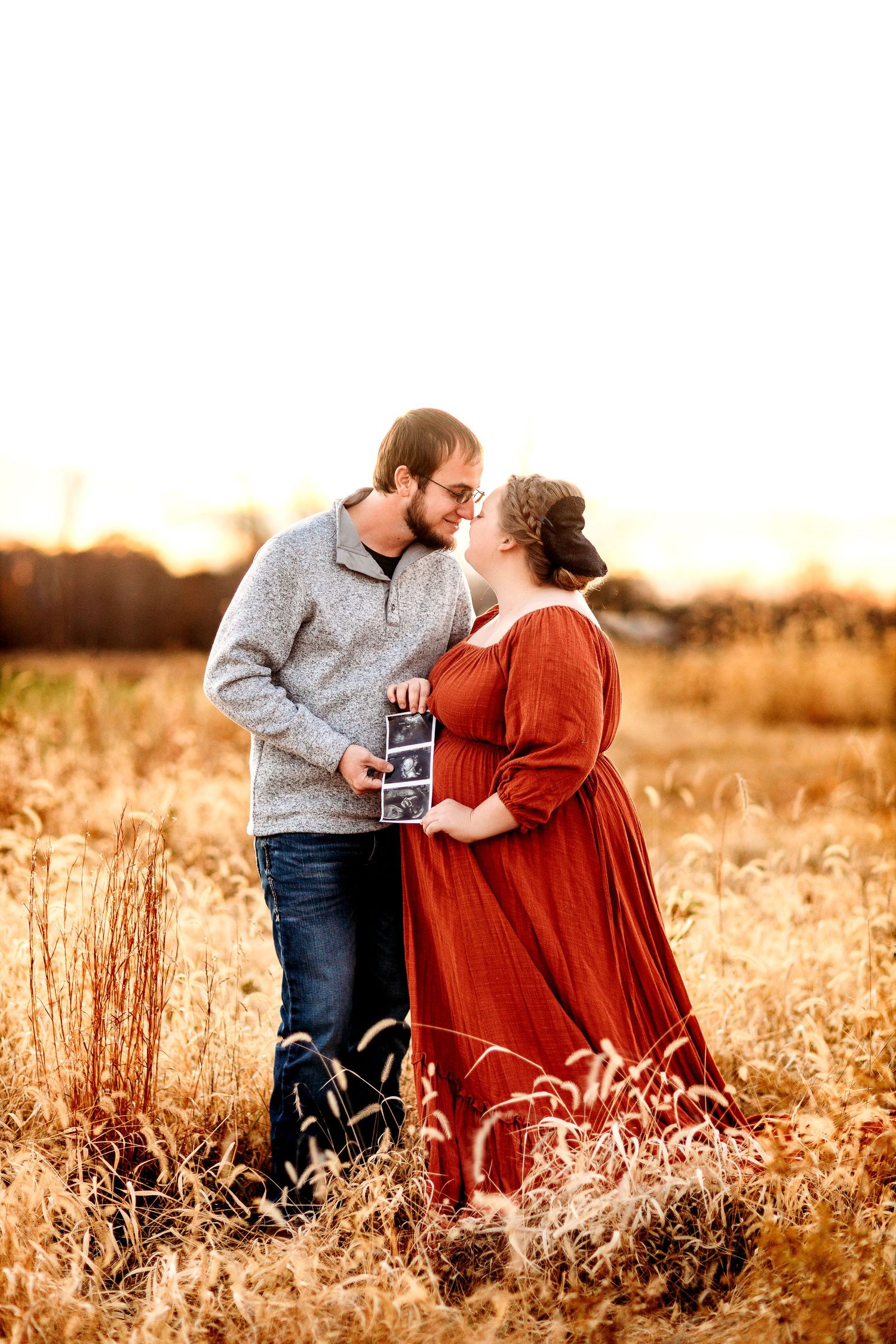 A couple stands in field at sunset, gently touching foreheads and smiling. They hold ultrasound photos. the woman wears a flowing red dress