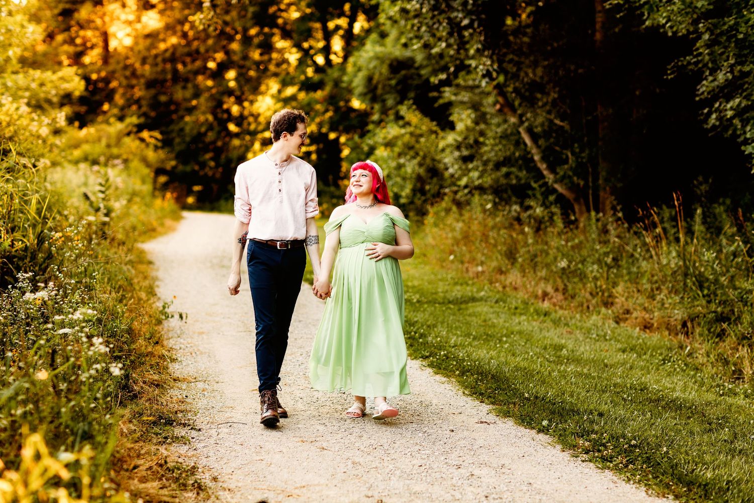 A couple walks hand in hand along a sunlit forest path. The woman in a light green dress smiles warmly at the man