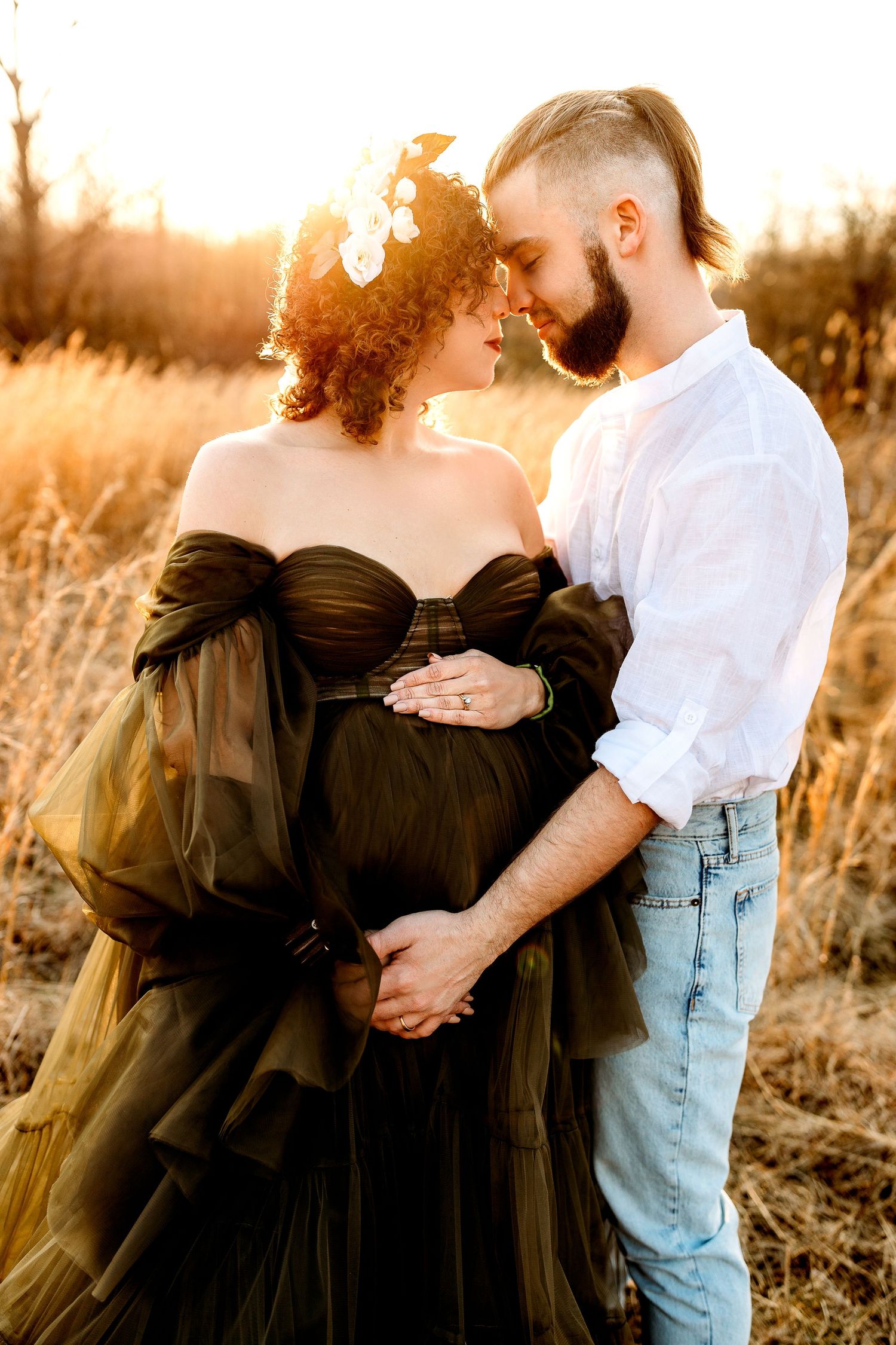 A couple embraces in a sunlit field. The woman wears a flowing gown, and the man in a white shirt gently holds her belly