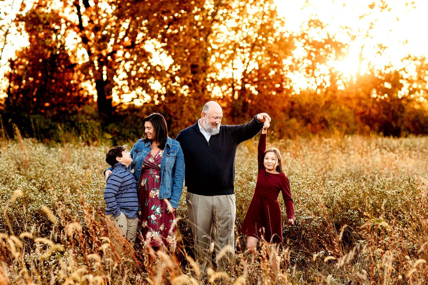 A family of four stands in a sunlit field. A man playfully twirls a girl by the hand, while a woman smiles at a boy. Warm autumn tones
