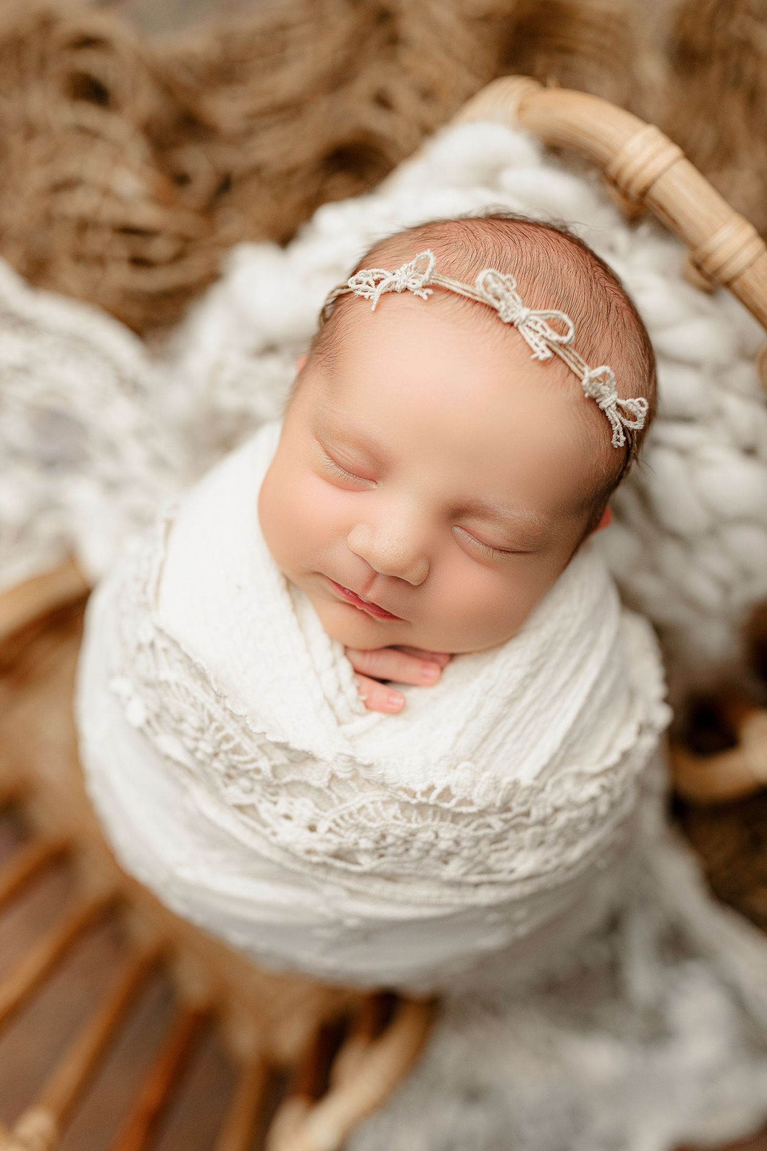 A newborn baby peacefully sleeps, swaddled in a white blanket. The baby wears a delicate lace headband.