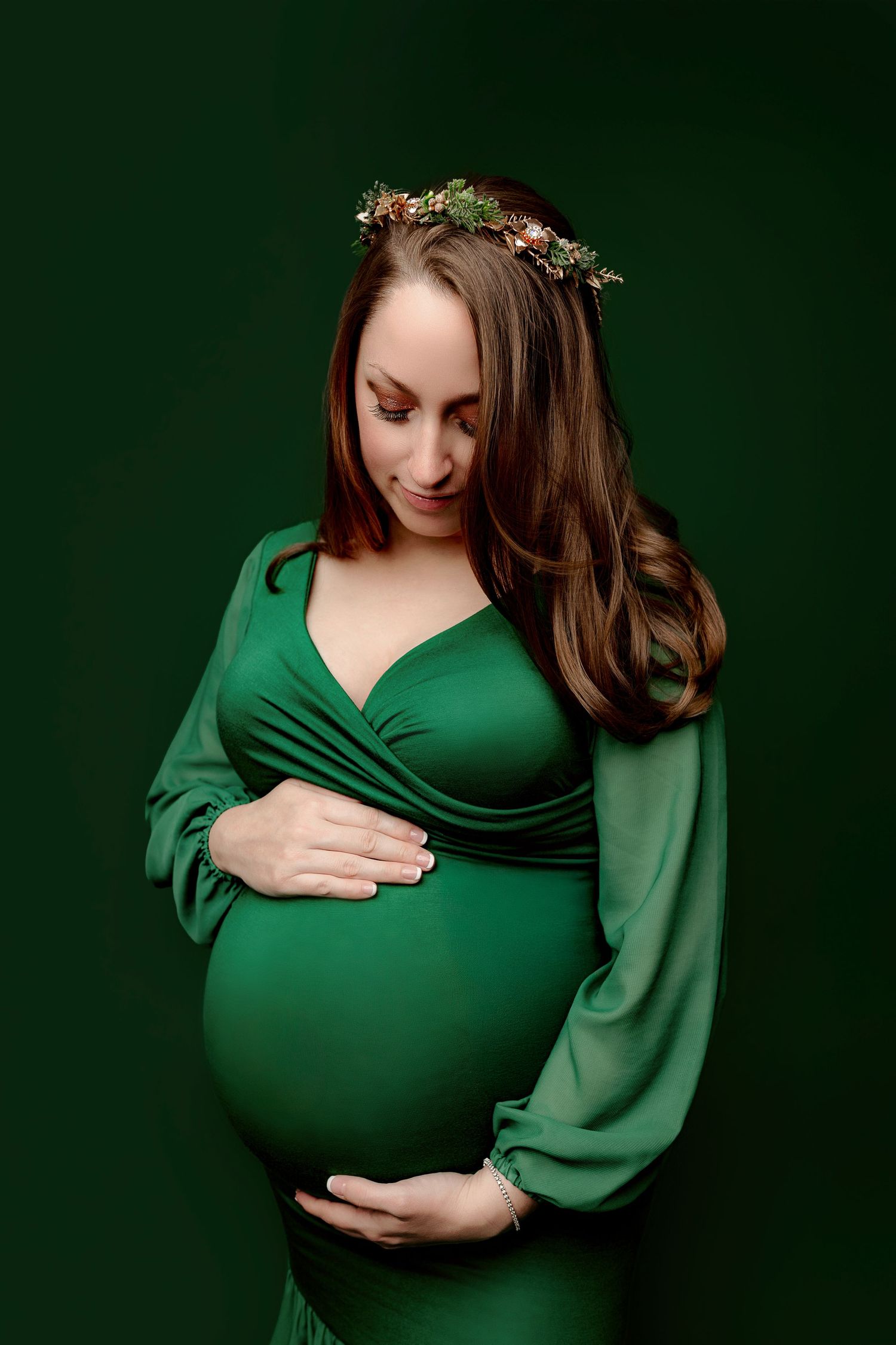 Pregnant woman in a green dress, gently cradling her baby bump. She wears a floral headpiece and looks down with a serene expression.