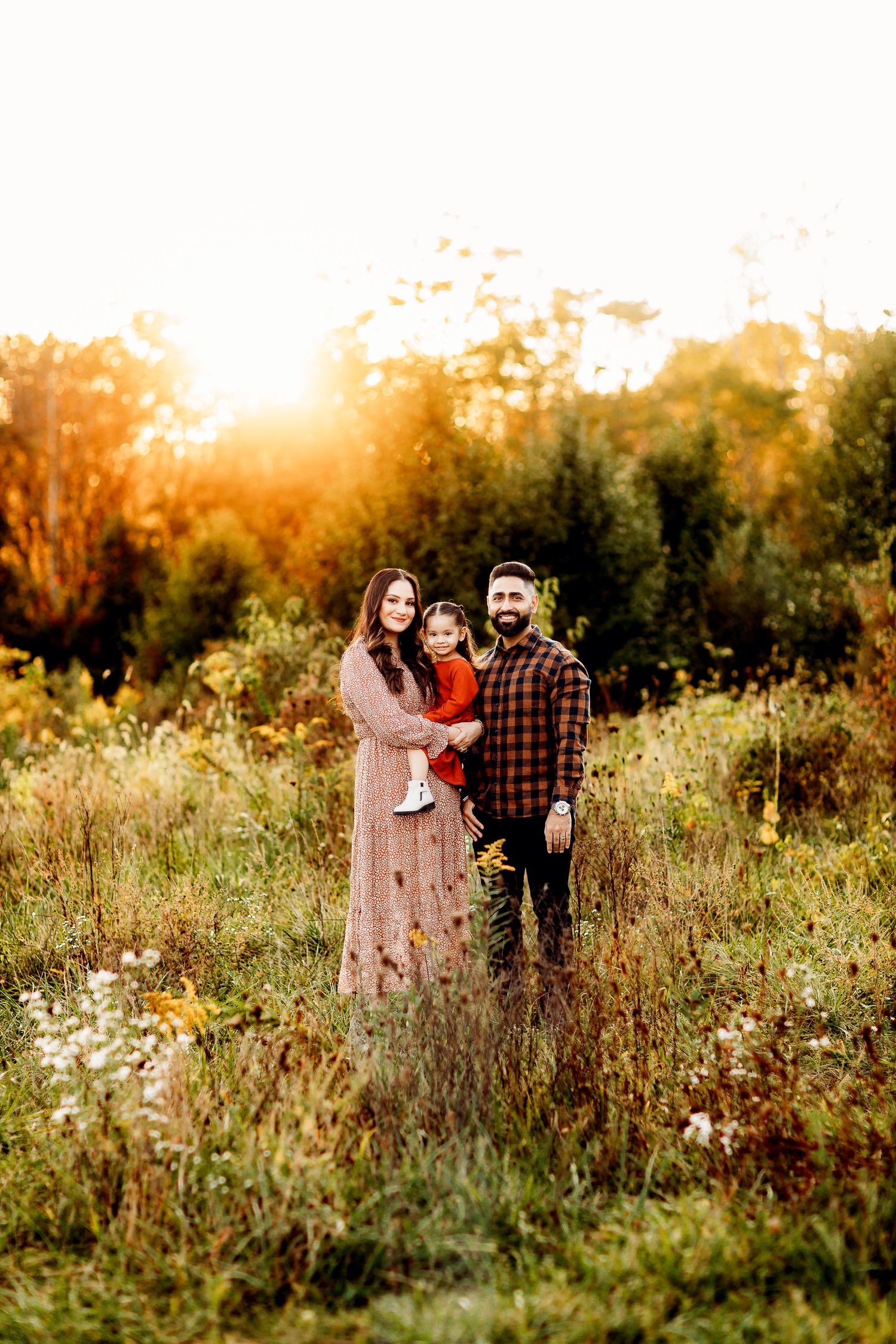 A family of three stands in a sunlit meadow. The woman wears a long dress, holding a child in red. The man is in a plaid shirt.