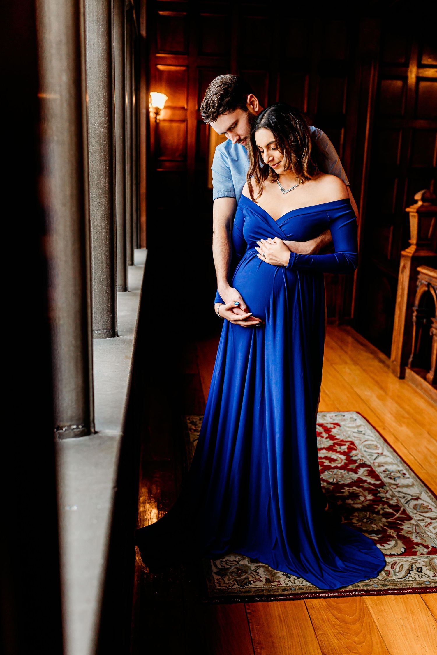 A couple stands in a dimly lit room by a window. The woman in a flowing blue dress cradles her baby bump, while the man gently embraces her.