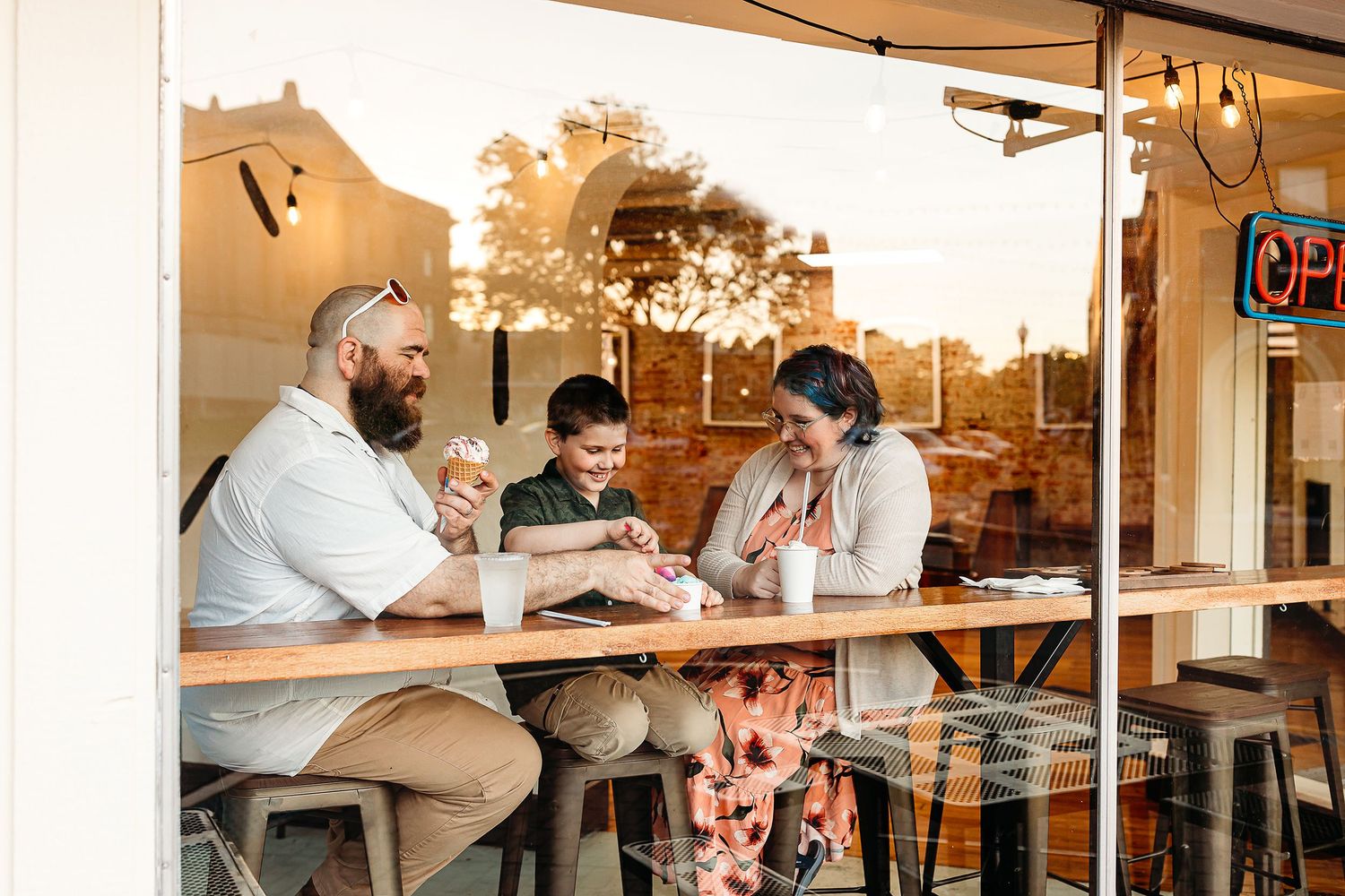 A family of three enjoys ice cream inside a cozy shop. They sit on stools by the window, smiling and conversing.