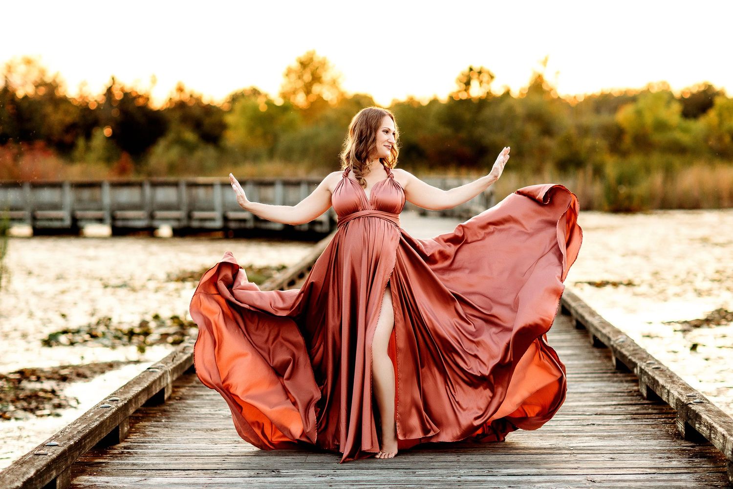 Woman in an elegant flowing rust-colored gown poses joyfully on a wooden path with a serene autumn landscape and soft sunset in the backgrou