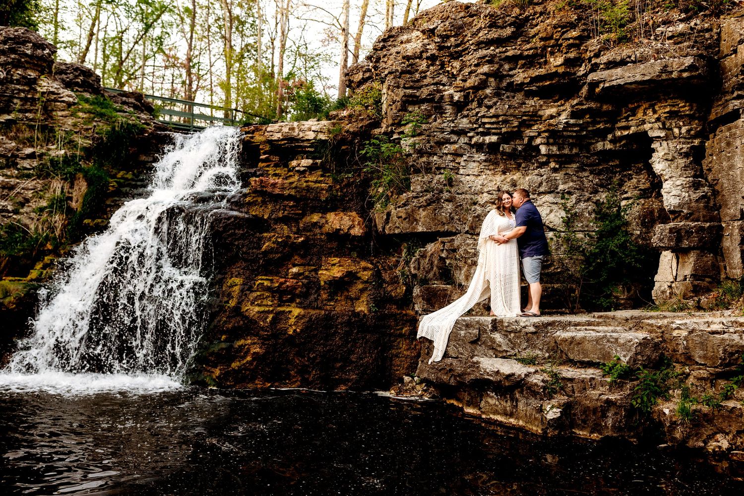 A couple embraces on rocky ledge by cascading waterfall, surrounded by lush greenery at France Park Indiana
