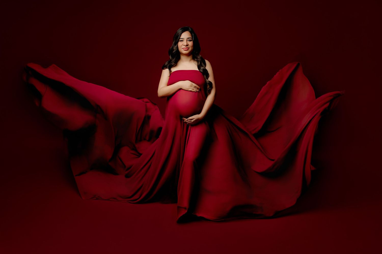 A pregnant woman in a flowing red gown poses against a matching red backdrop in Indianapolis Maternity Photography Studio