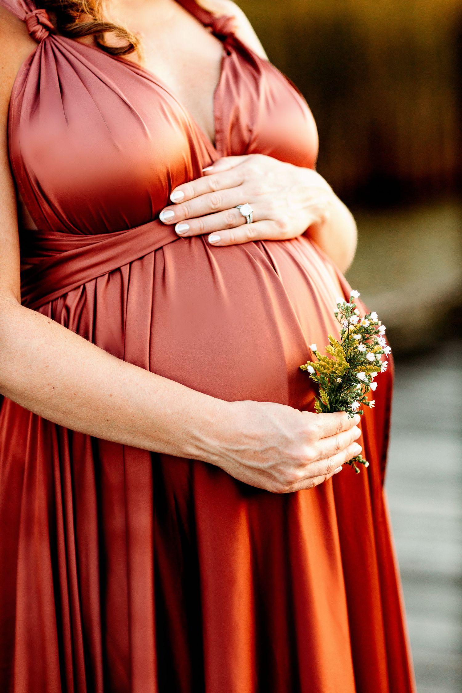 A pregnant woman in a flowing rust-colored dress gently cradles her belly with one hand and holds wildflowers with the other