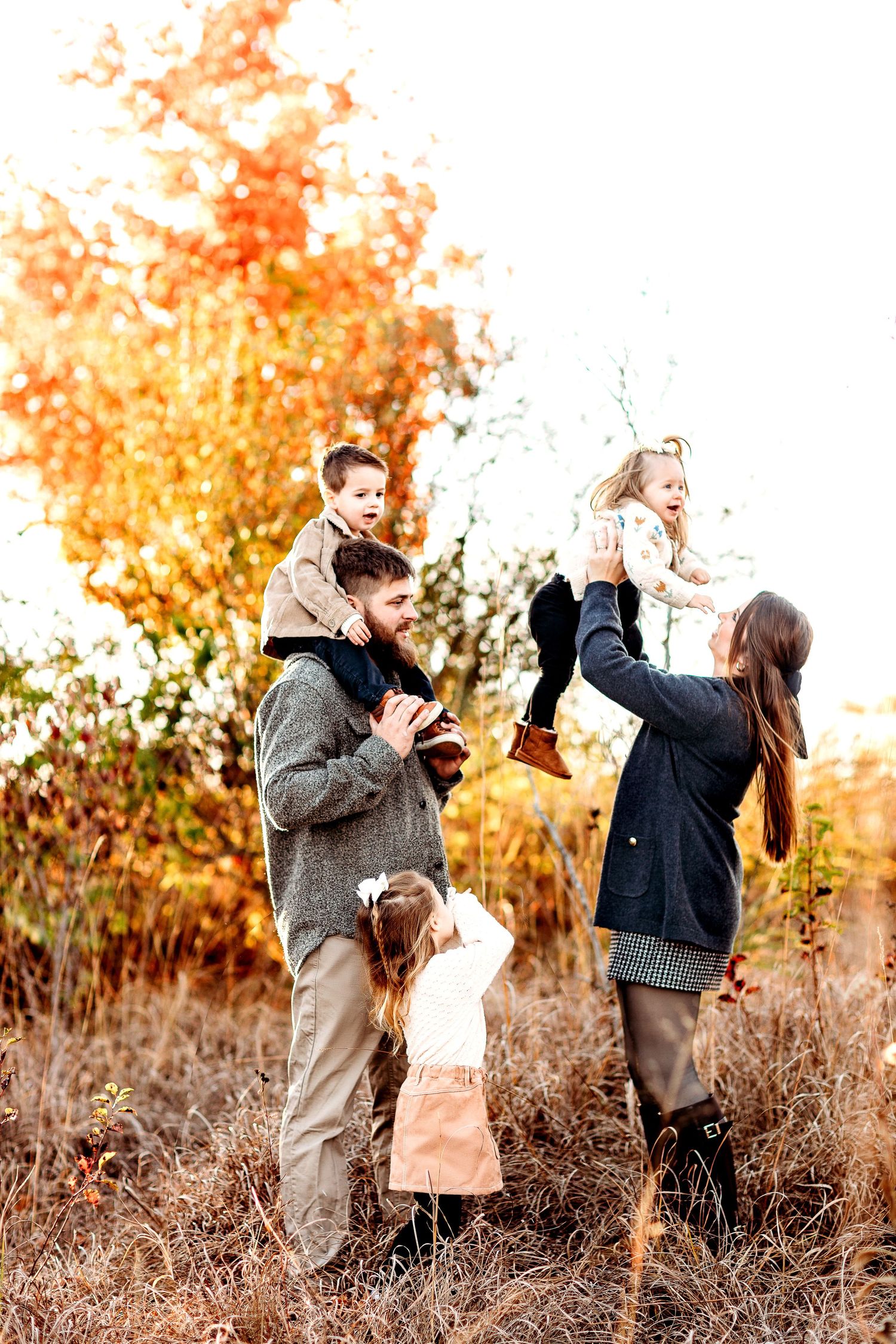 A family of four enjoying an autumn day in a field, with vibrant fall foliage. Parents lifting and playing with children, conveying warmth