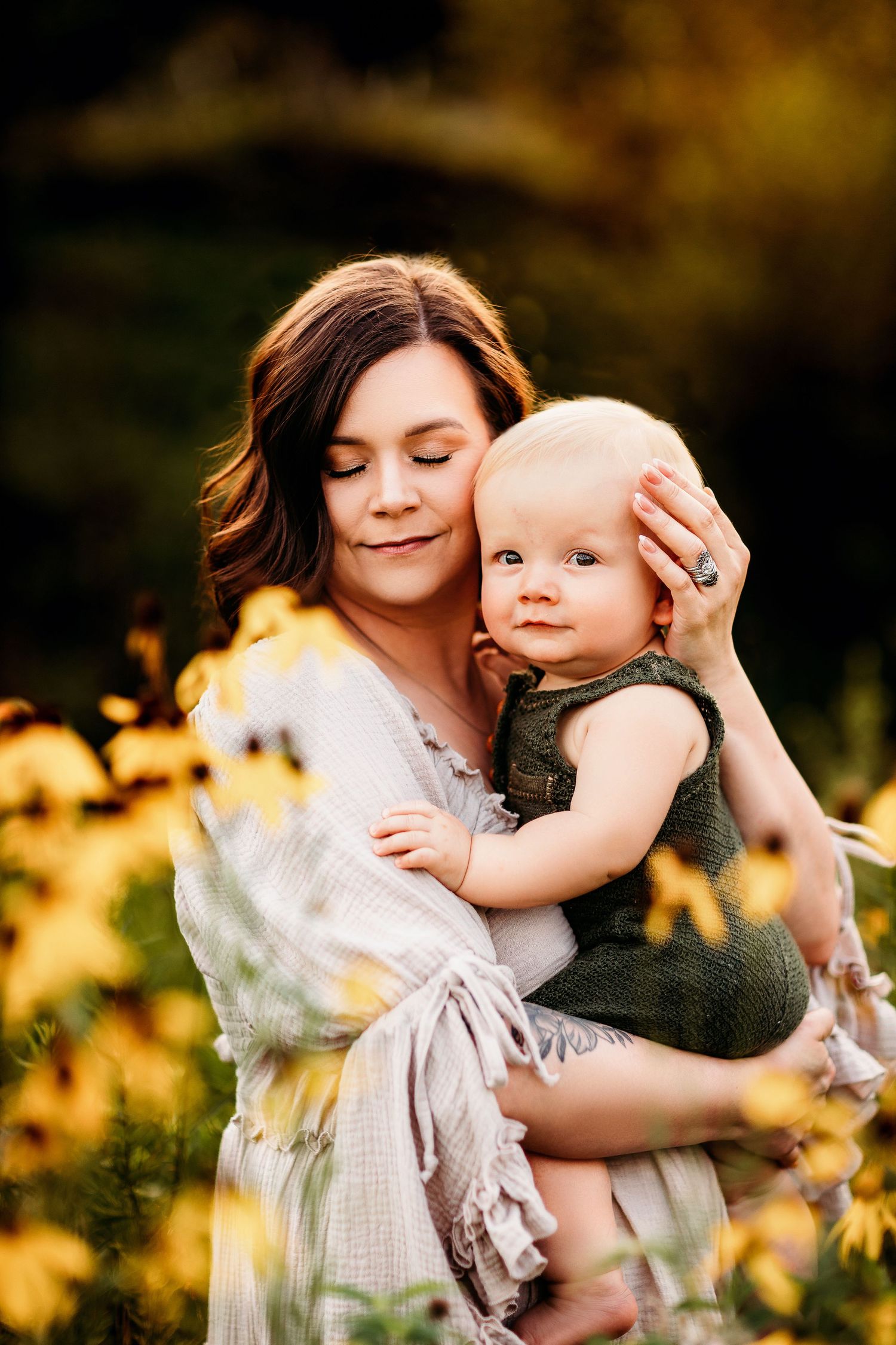 A woman gently holds a baby in a field of yellow flowers, both with serene expressions. The warm sunlight creates a soft, golden glow