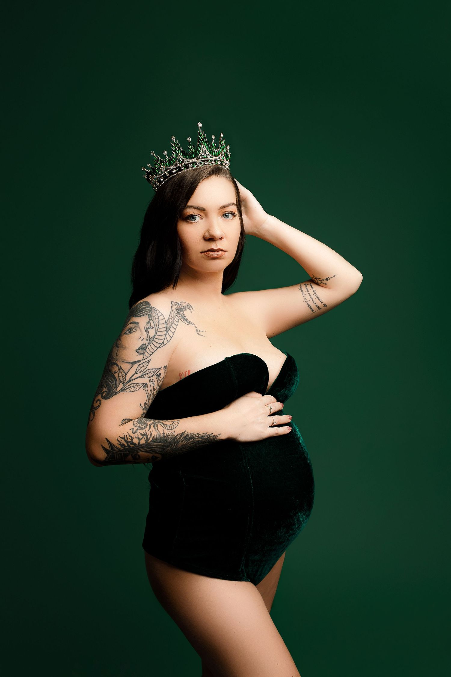 Pregnant woman in a strapless green gown and crown poses confidently against a green backdrop, hand on belly, showcasing tattoos