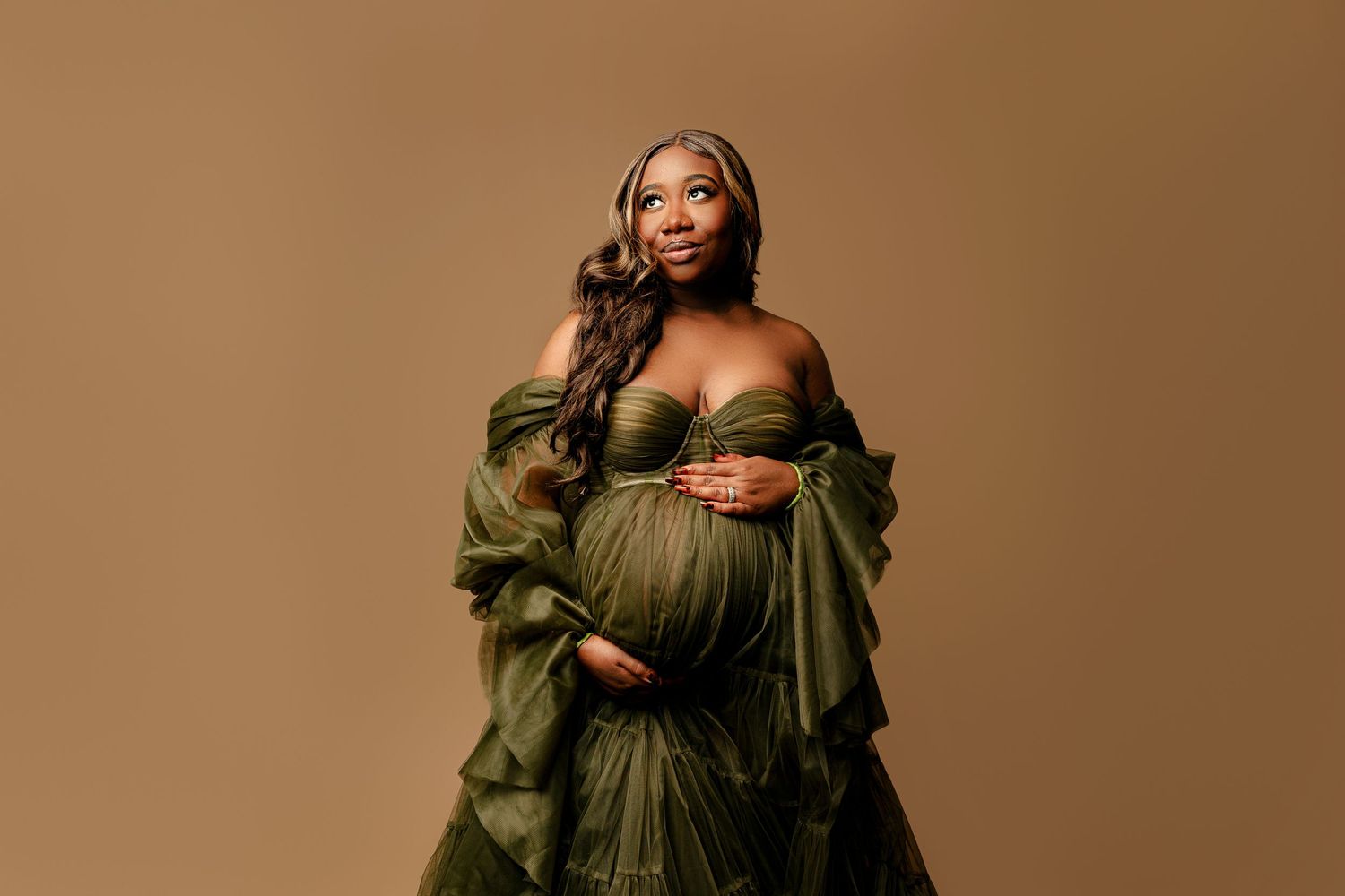 Pregnant woman in an elegant off-shoulder olive gown cradles her belly, standing against a soft brown background. She gazes upwards