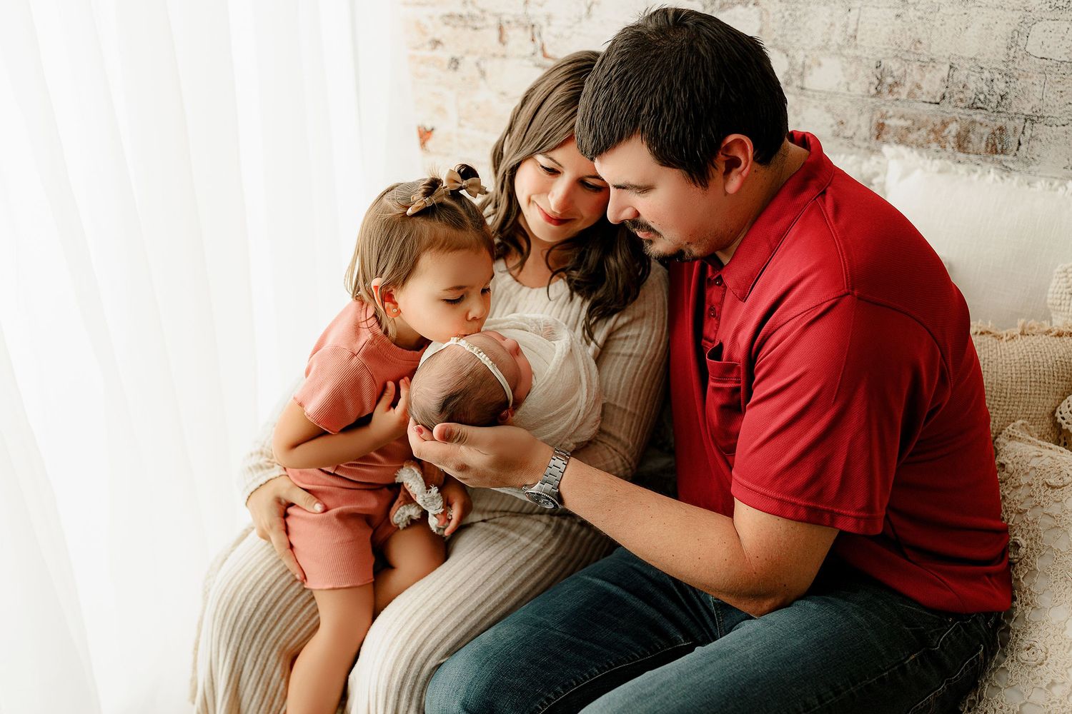 A family of four sits closely together on a sofa, with the father in a red shirt, mother in a striped dress holding a newborn, and a toddler