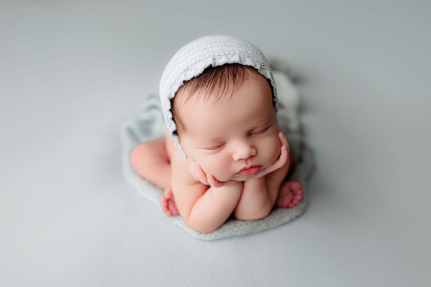 A peaceful newborn baby lies on a soft, light blue blanket, wearing a white knit bonnet, hands under chin timeless newborn portraits Indiana
