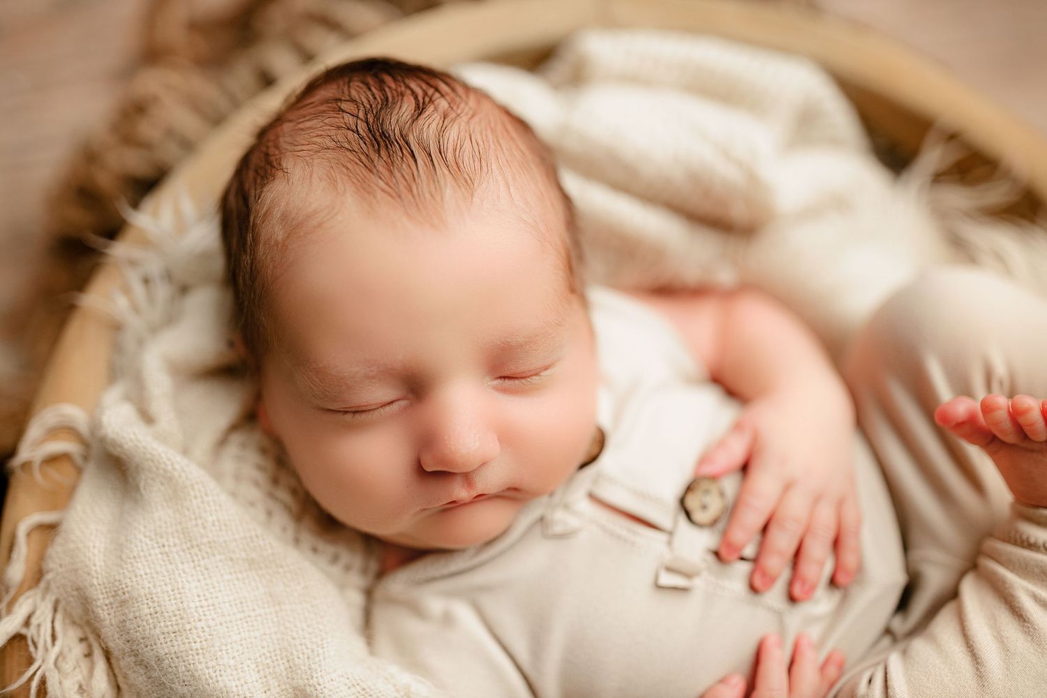 A sleeping newborn baby lies peacefully in a soft cream blanket inside a wooden basket. The baby's serene expression conveys warmth