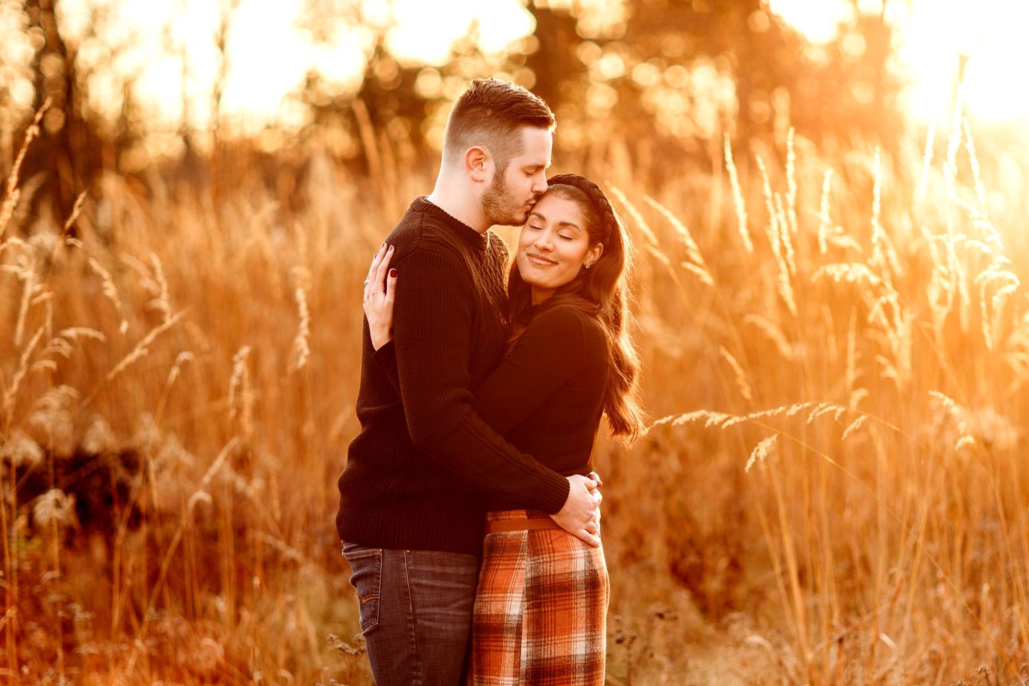 A couple embraces in a sunlit field. The man kisses the woman's forehead as they stand among tall grasses